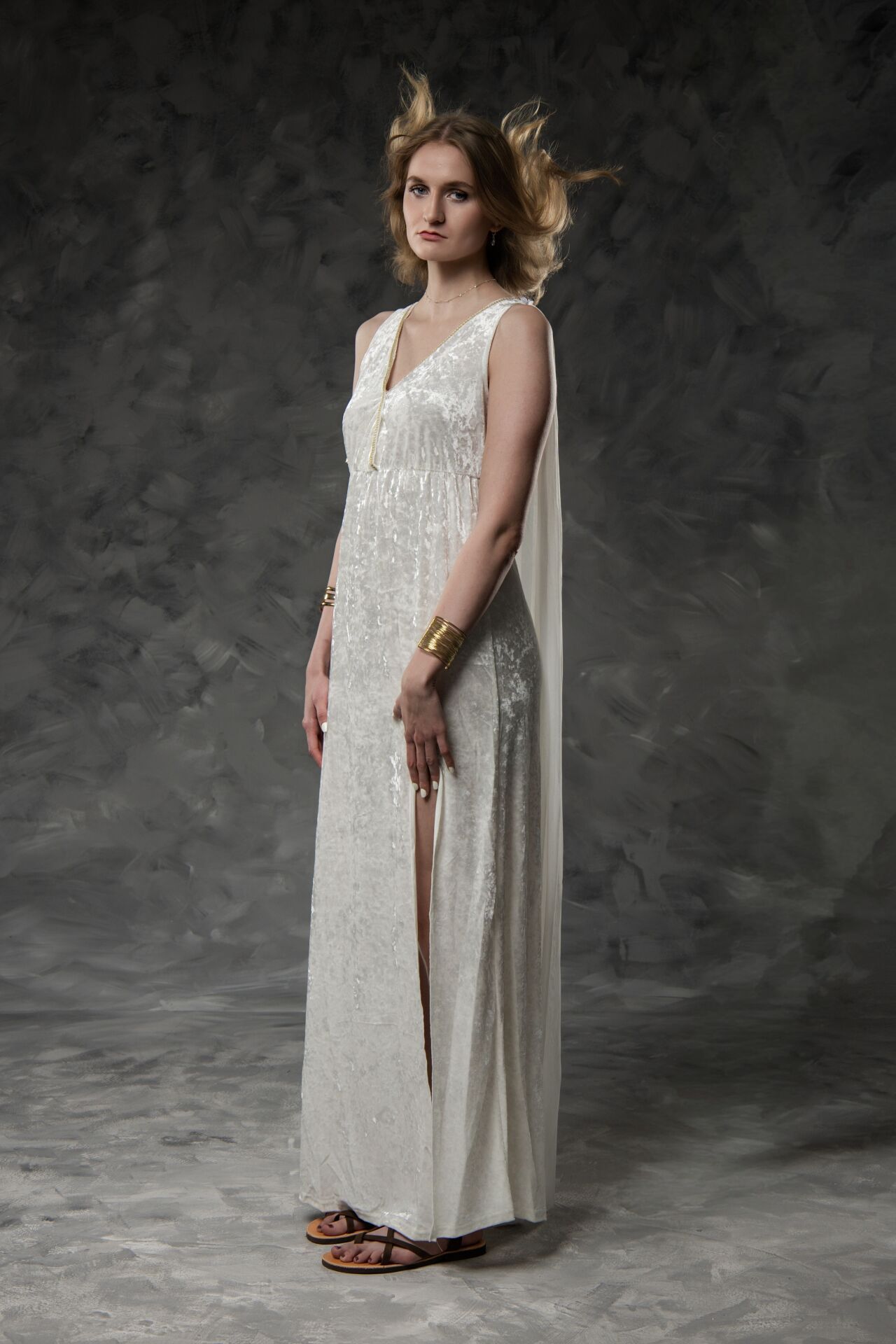 Woman in white grecian gown with gold cuffs and sandals poses against gray backdrop.