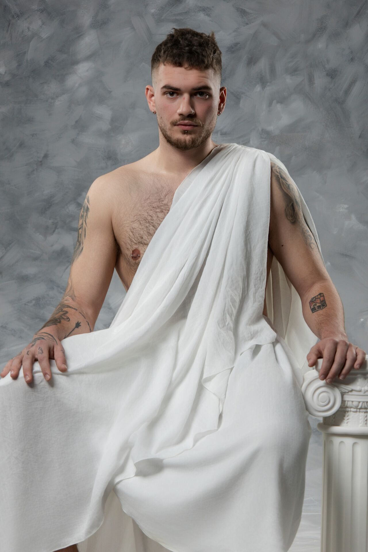 Classical style photoshoot featuring a figure in white toga-like drapery against a mottled grey background.