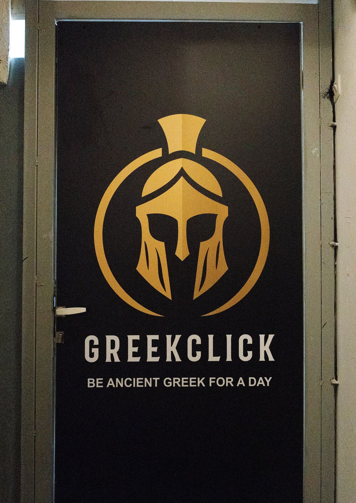 Black wall display showing gold Greek warrior helmet logo and text 'GREEKCLICK - BE ANCIENT GREEK FOR A DAY'.