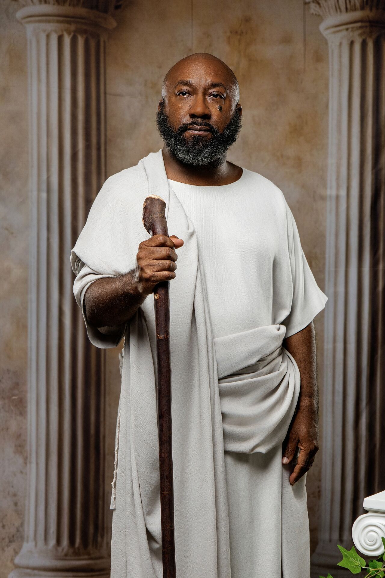 Bald Black man with gray beard in white robe holding wooden staff, standing between stone columns.