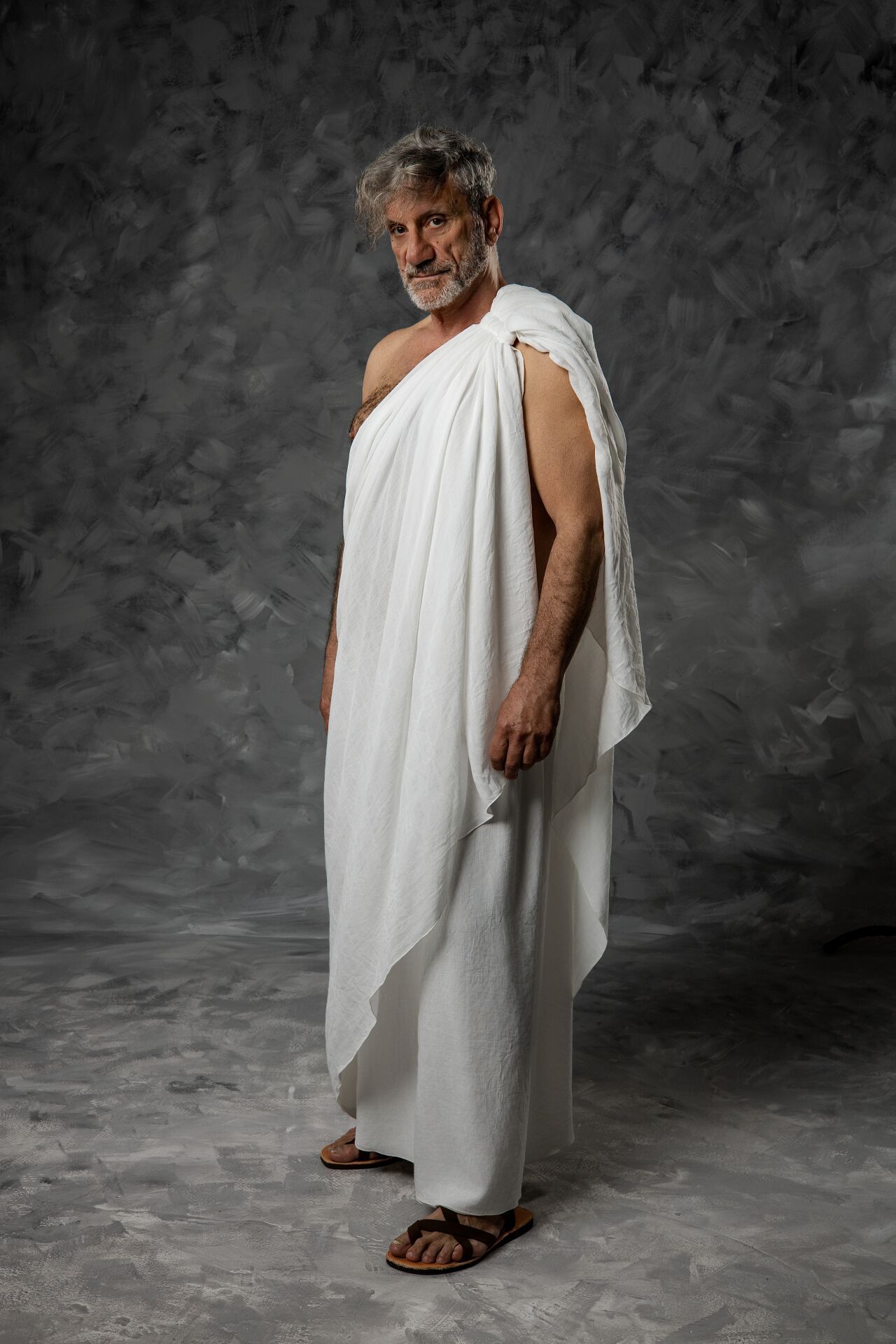 Elderly man in white Roman toga and sandals poses against gray textured background in classical ancient Greek style.