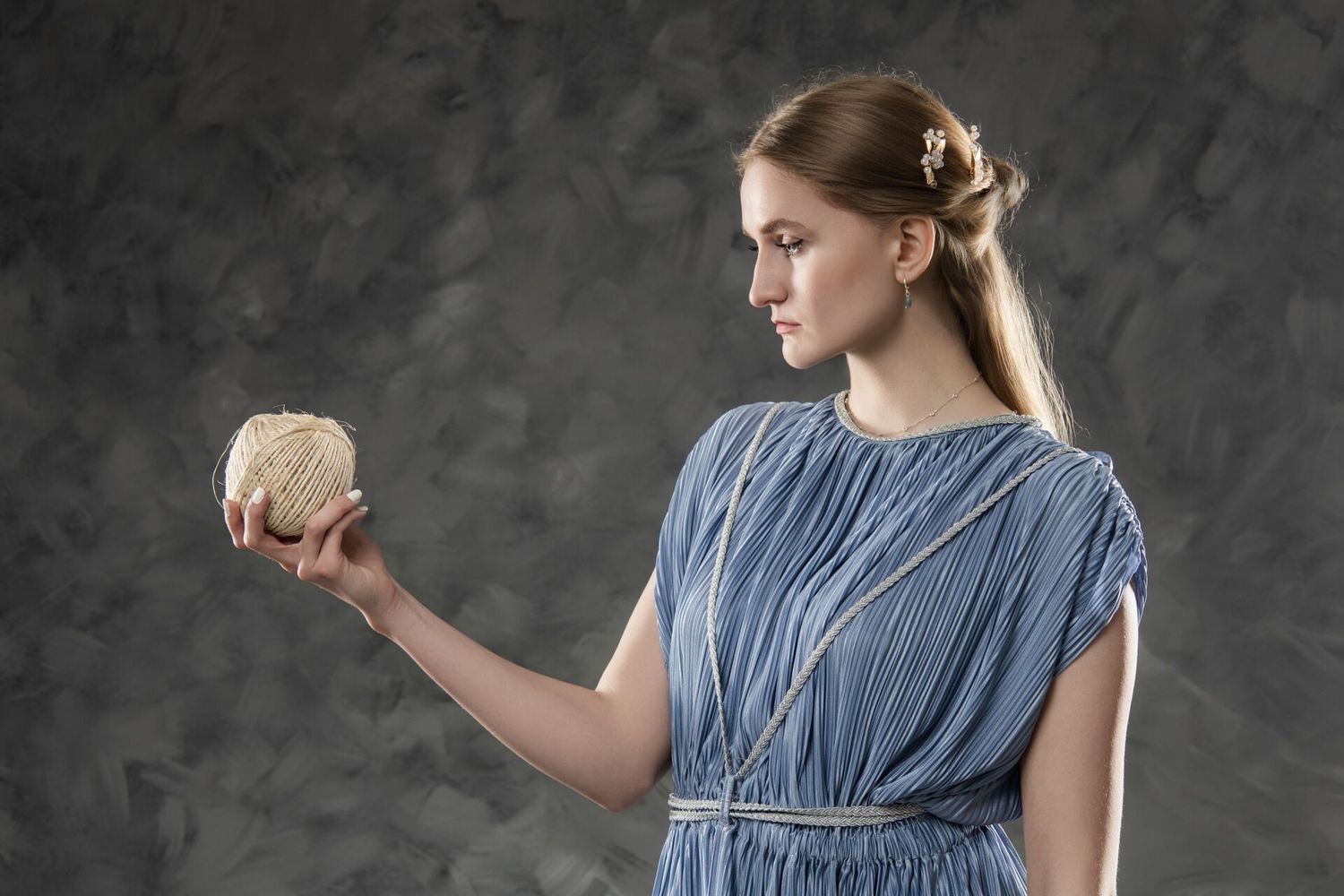 Young woman in blue Greek dress holding a decorative golden sphere against a gray textured background.