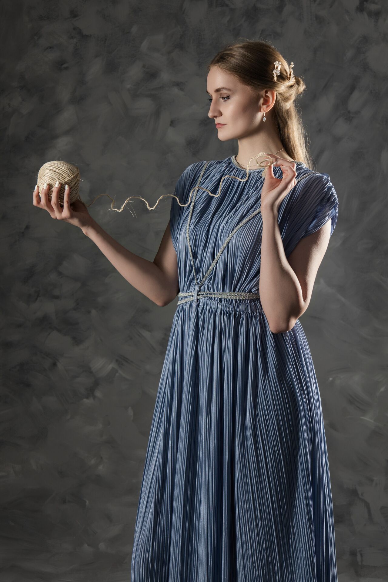 Elegant woman in blue pleated dress holding a ball of twine against gray textured background.