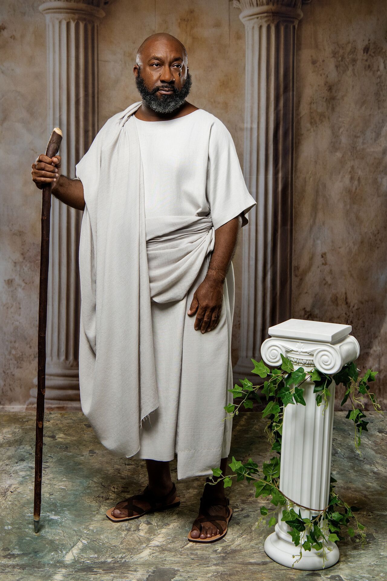 Homer holding a wooden staff beside an ivy-covered column on a marble floor.