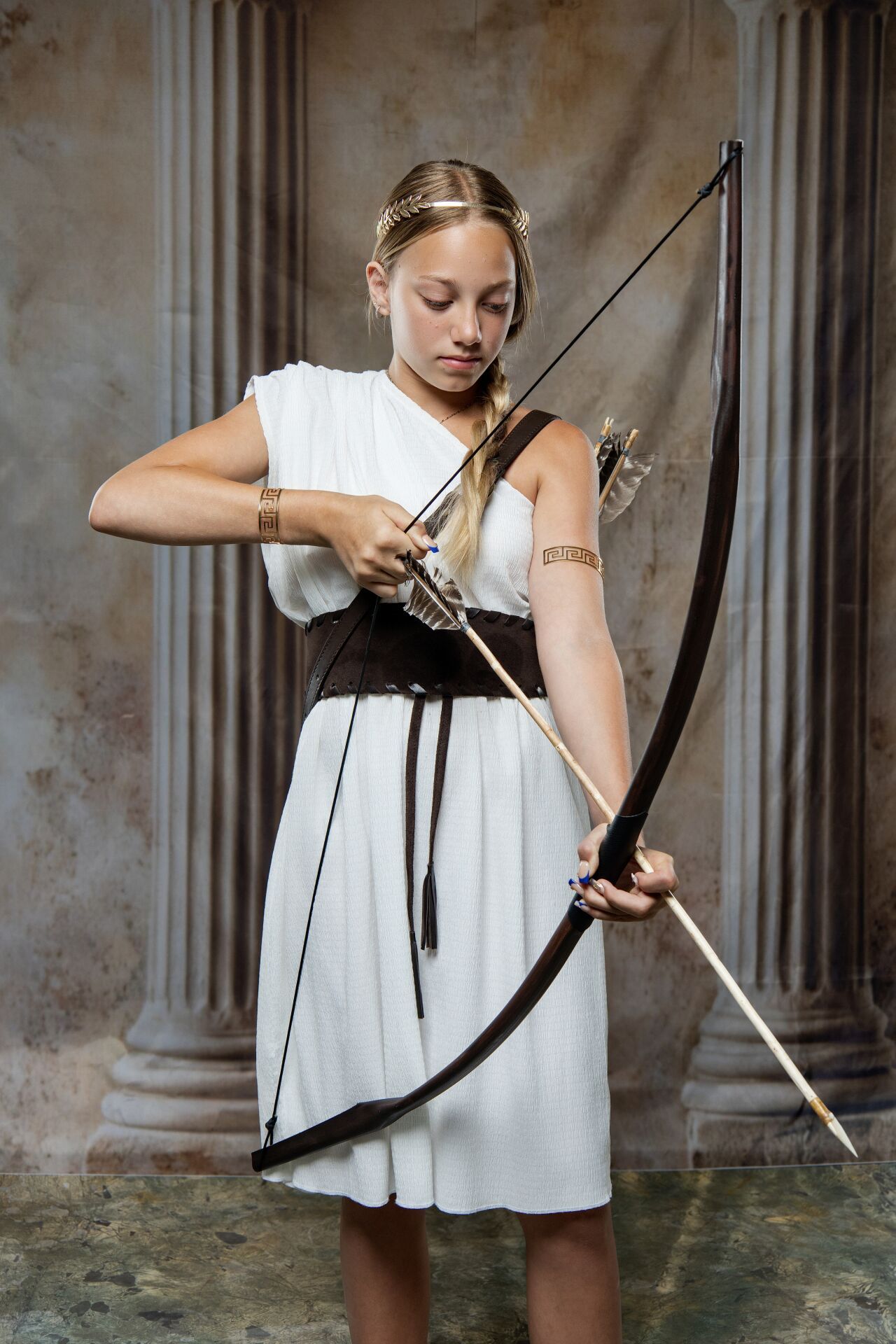 Young woman in white Greek dress nocks arrow on longbow, wearing gold arm cuffs before stone columns.