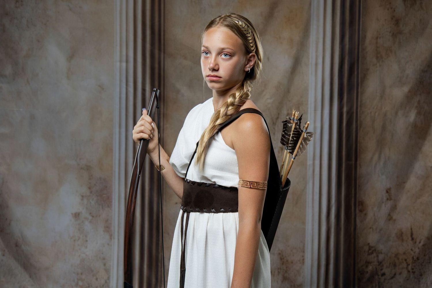 Young woman in white Greek goddess costume with braided hair, holding bow with quiver of arrows on back.