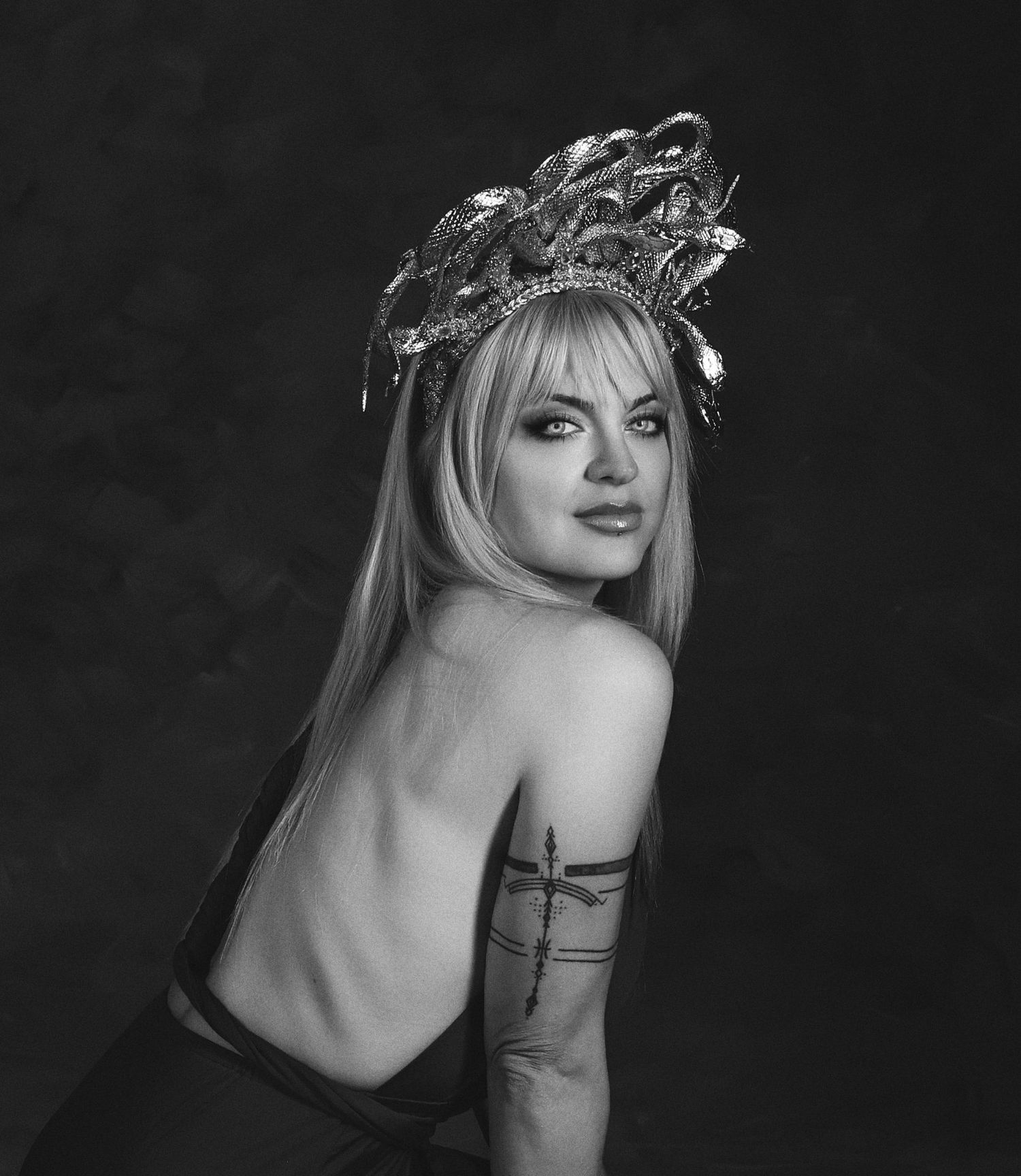 Black and white ancient Greek-inspired portrait of person wearing ornate laurel crown headpiece, bare shoulder with arm tattoos, artistic