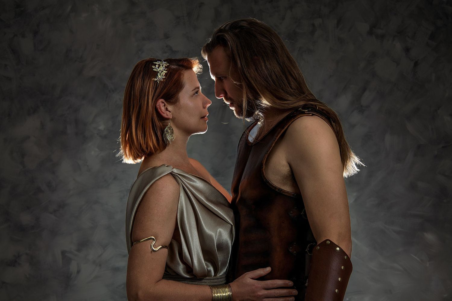 Two people in ancient Roman costumes share an intimate moment, facing each other closely in a dramatic studio setting.