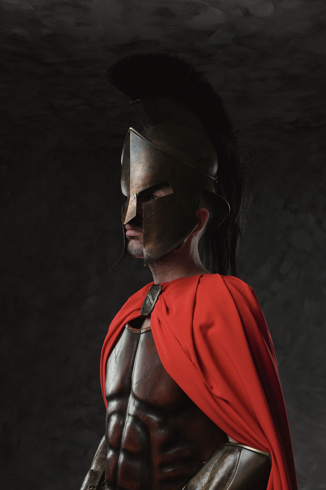 Dramatic studio portrait of a muscular figure wearing a flowing red cape against a dark background.