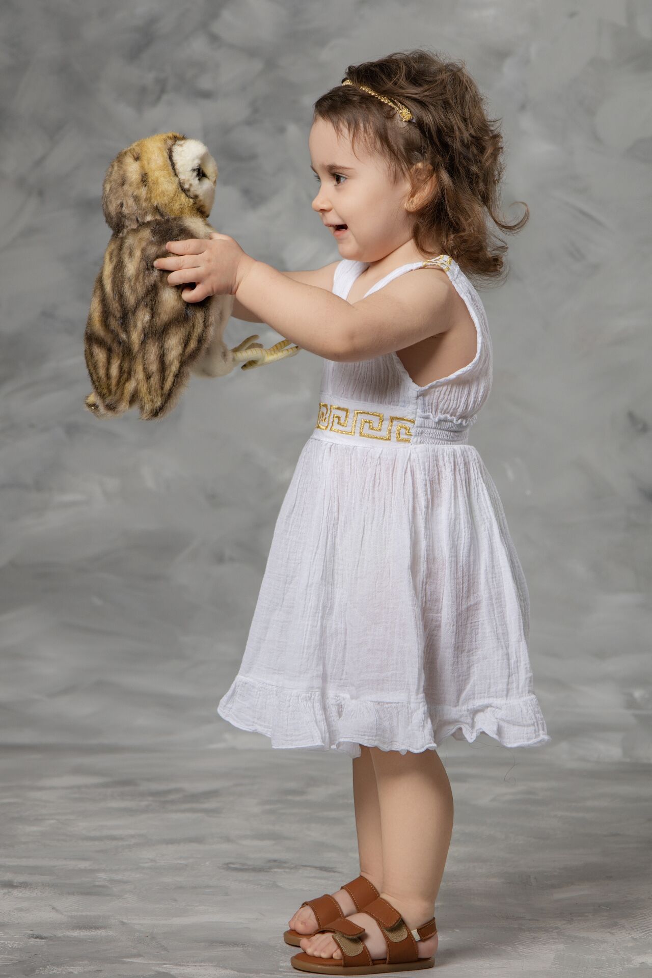 A child in a white dress and brown sandals holds a small stuffed animal while standing against a gray background.