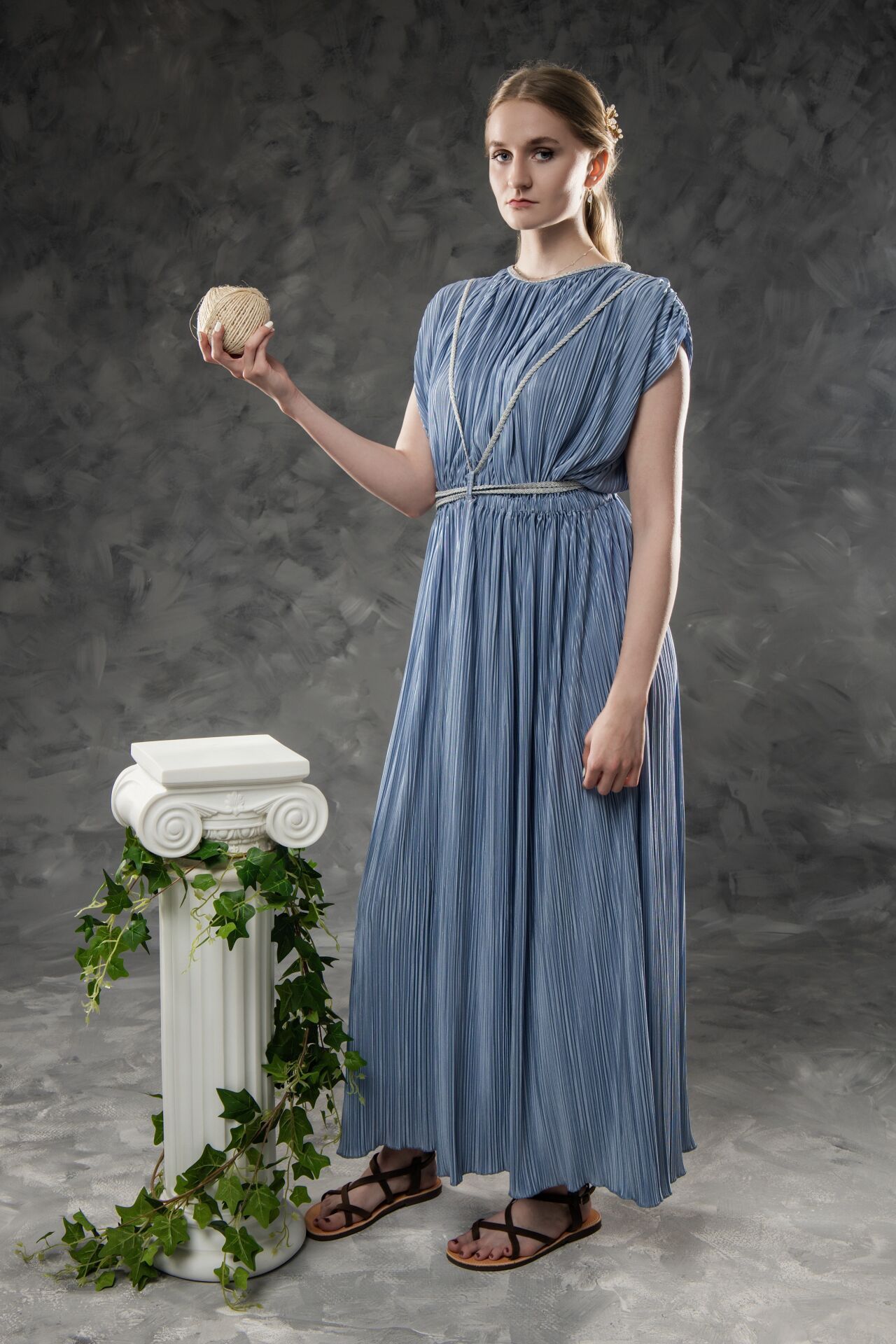 Young woman in blue Greek-style pleated dress holding a skull beside an ivy-draped white column on gray background.
