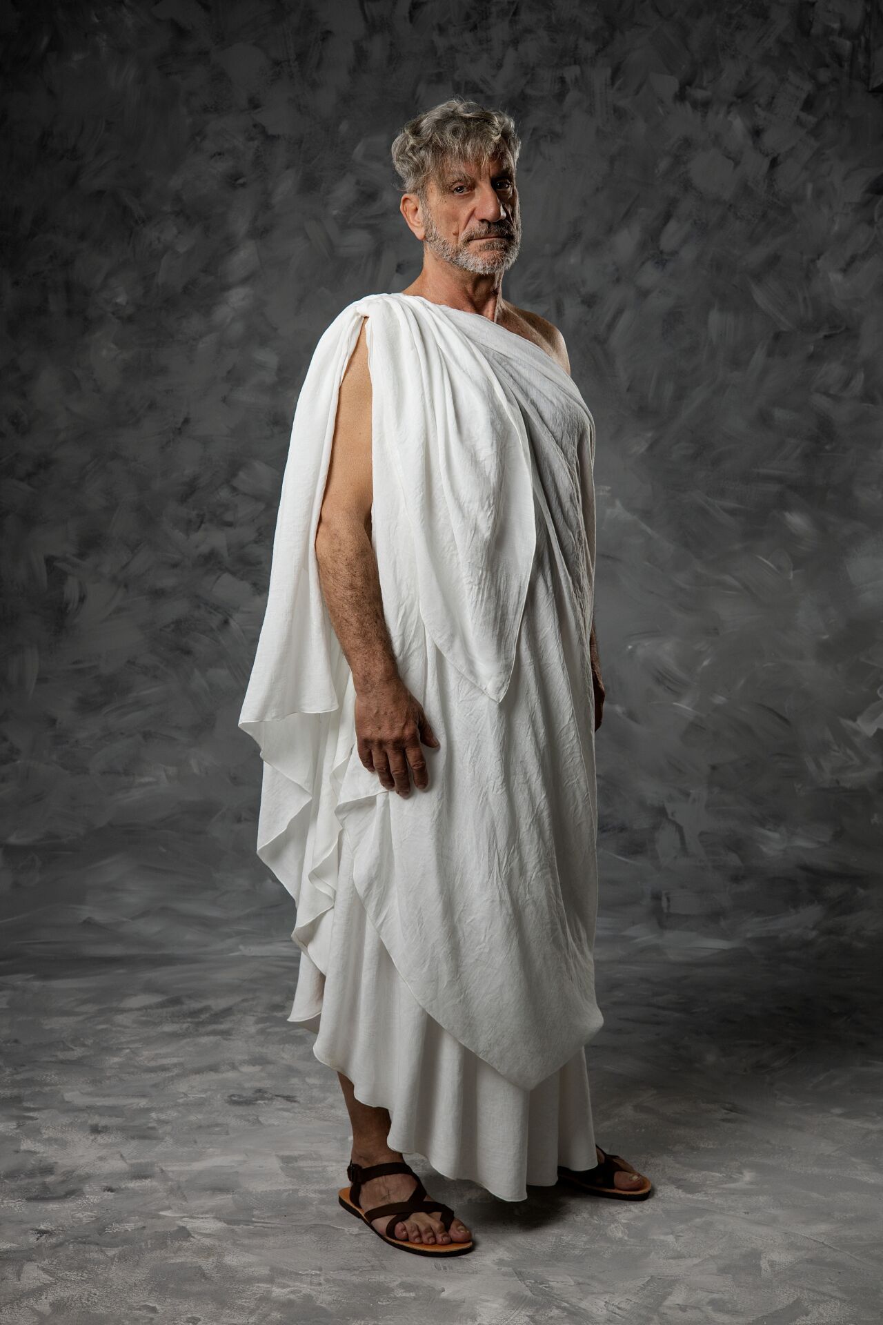 Middle-aged man in ancient Greek white toga and sandals posing against gray textured studio background.