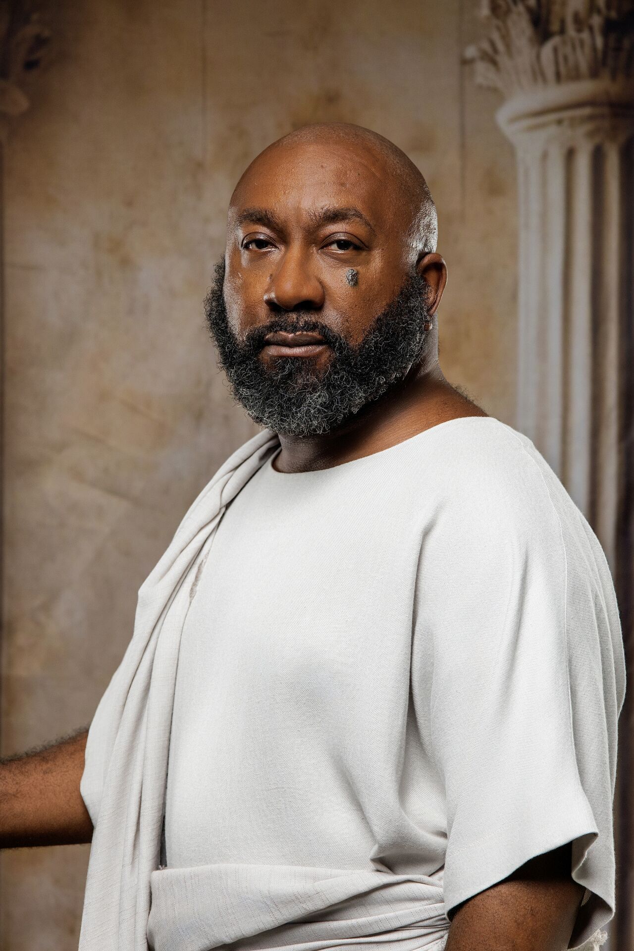 Bald Black man with gray beard wearing white toga, posing before marble column background.