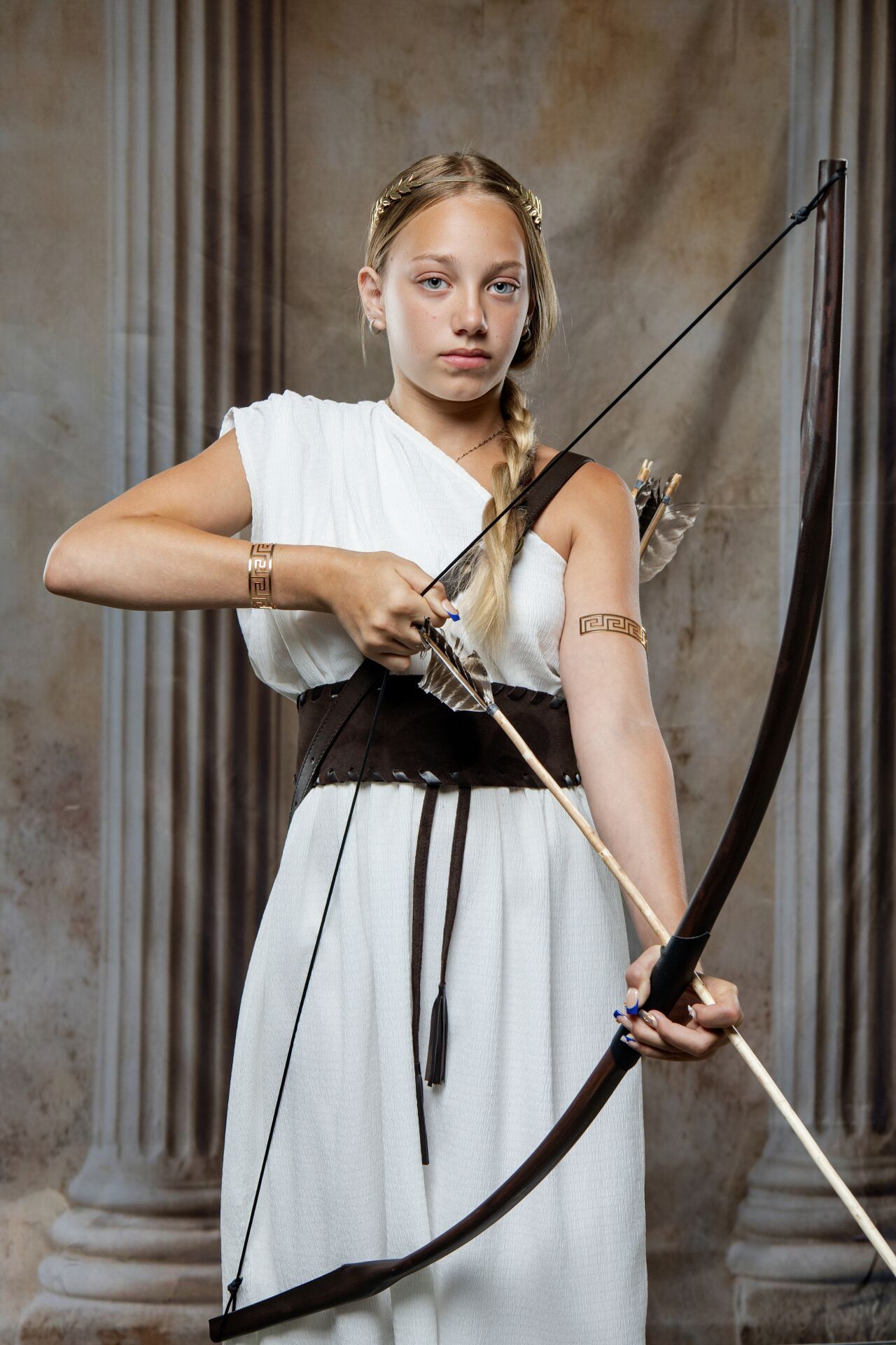 Young woman in white Greek goddess costume drawing a bow and arrow, wearing gold armband and dark leather belt.