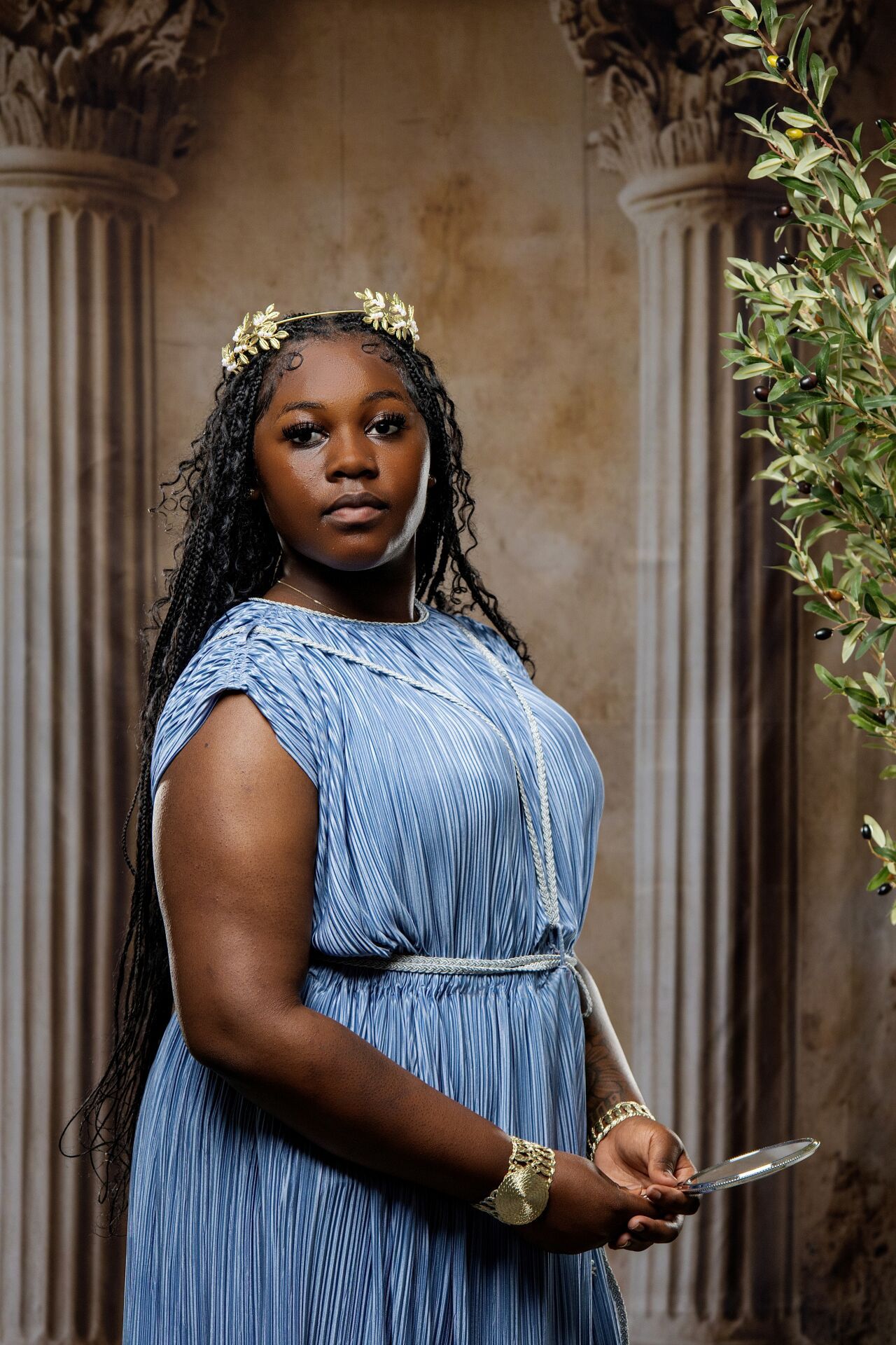 Young Black woman in blue Greek-style dress with gold crown and bracelet poses before ancient columns.
