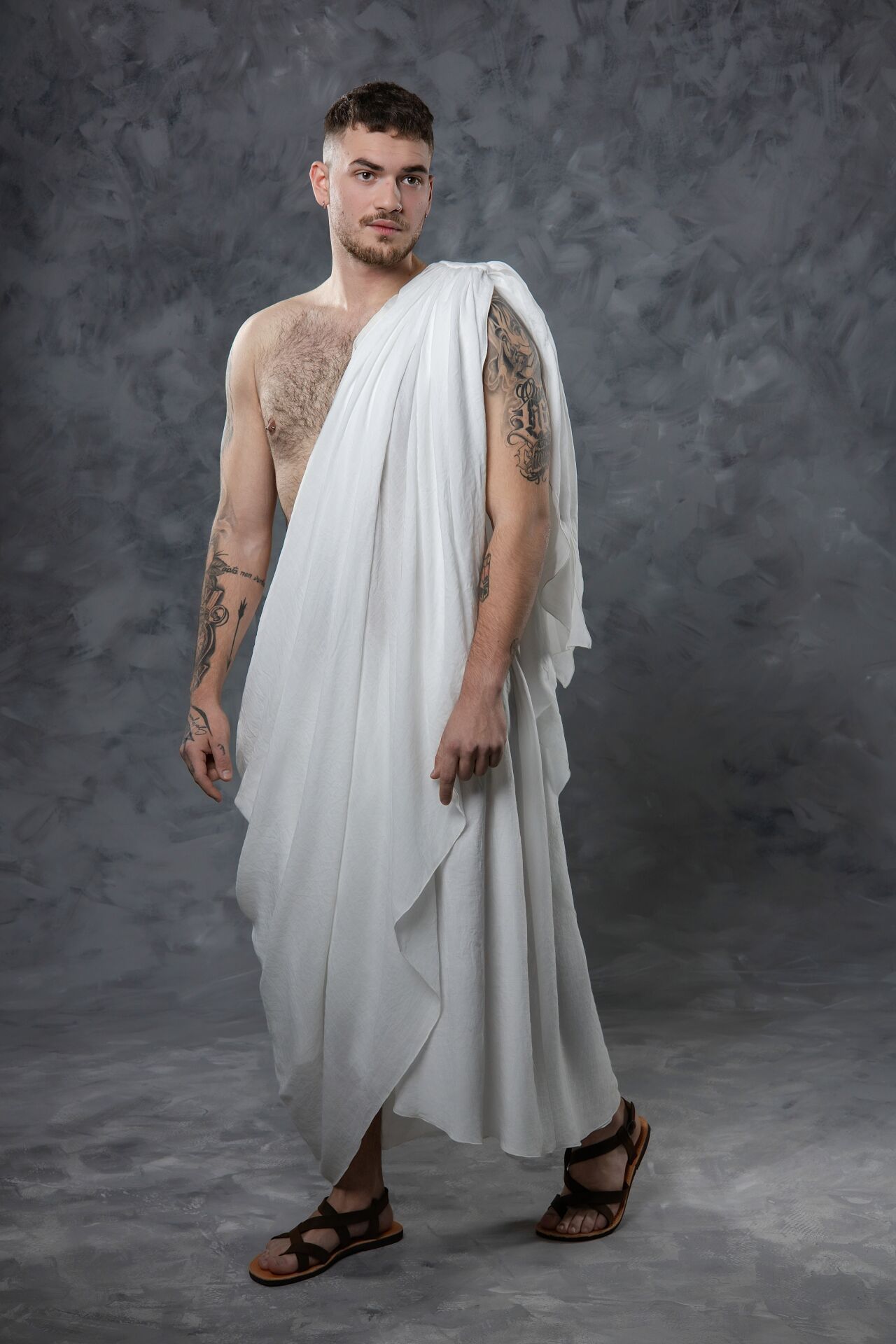 A person wearing a flowing white toga-style garment and brown sandals poses against a gray backdrop in multiple poses.