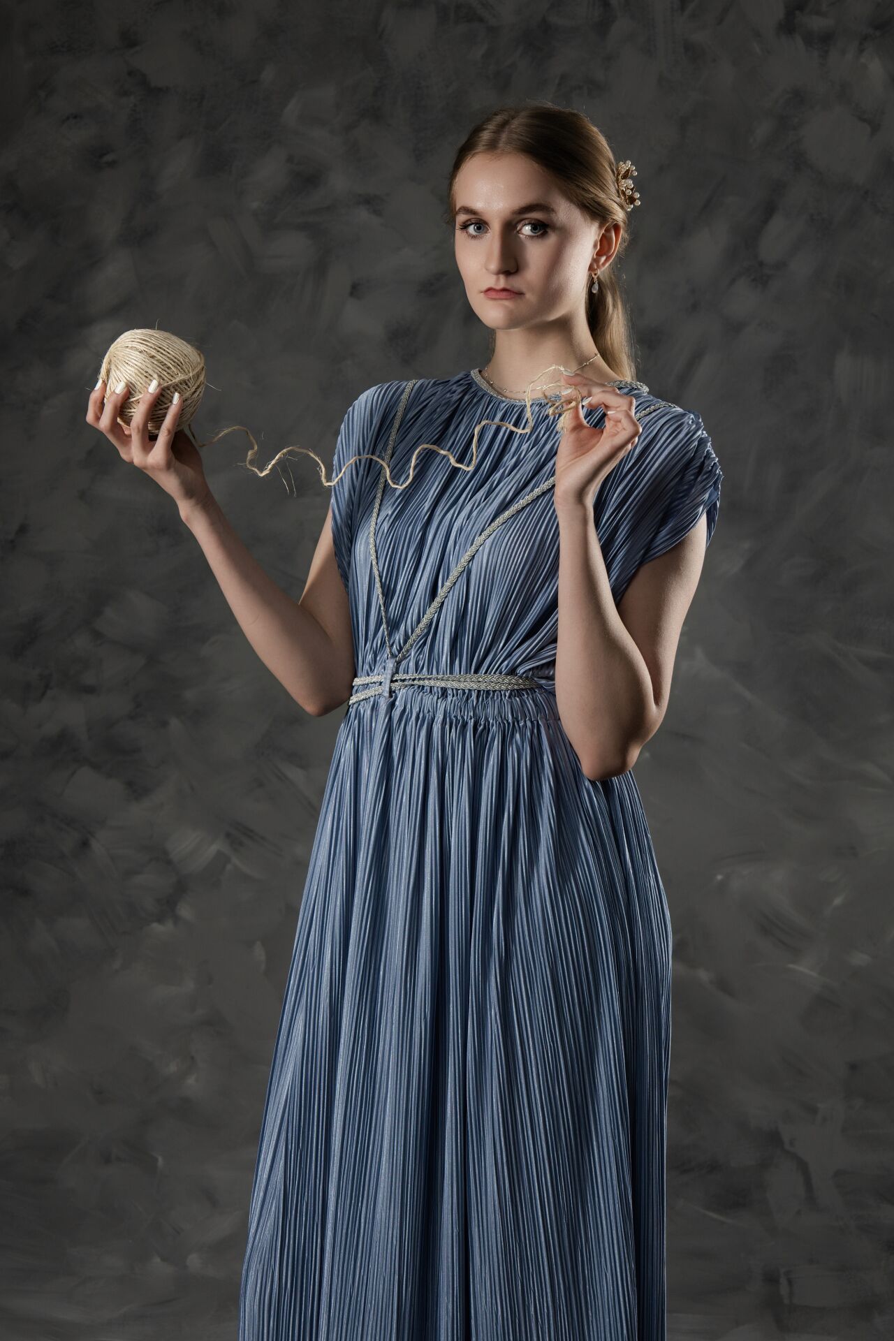 Young woman in blue pleated dress holds golden ball of twine against dark textured background.