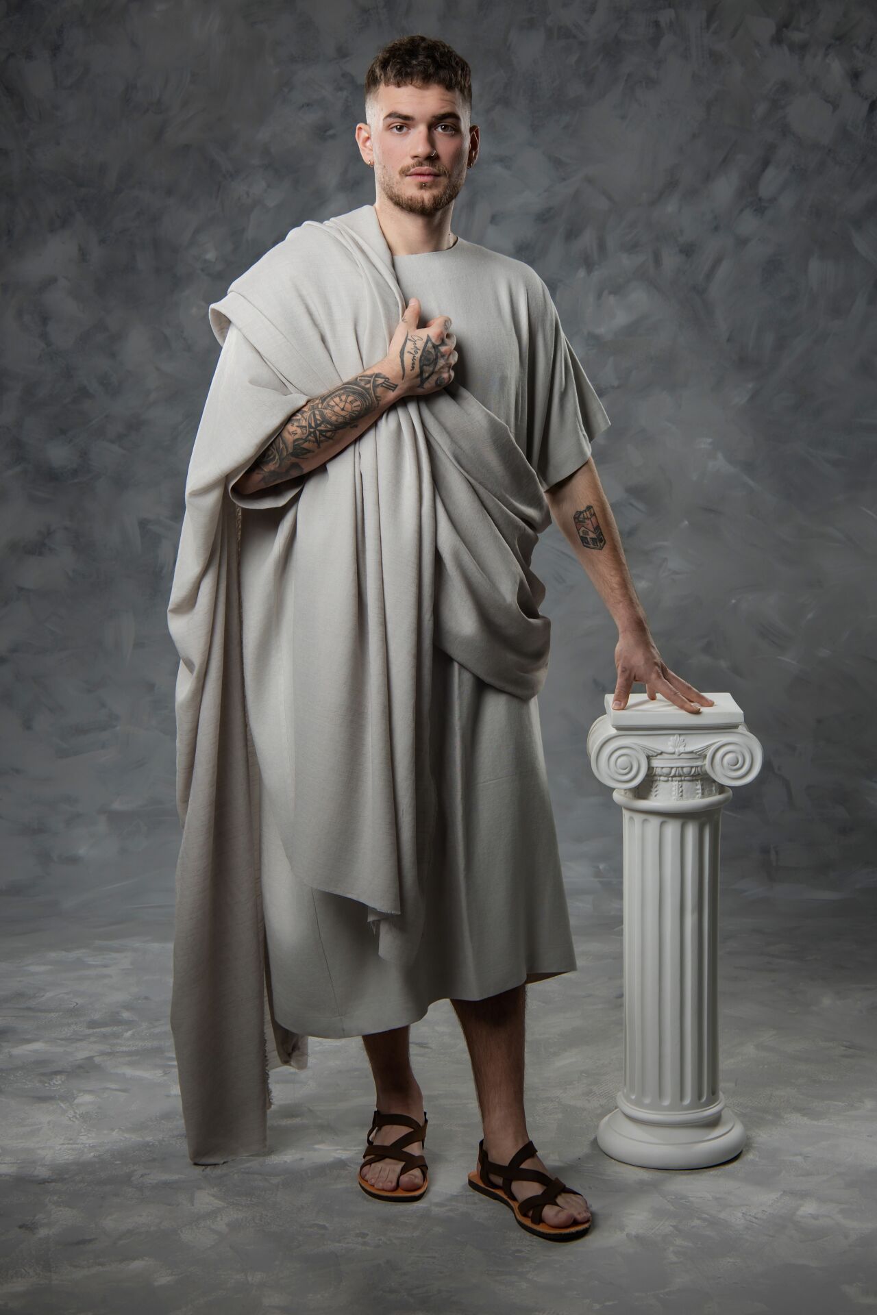 Person wearing flowing beige toga-style garment poses next to decorative white classical column in studio setting.