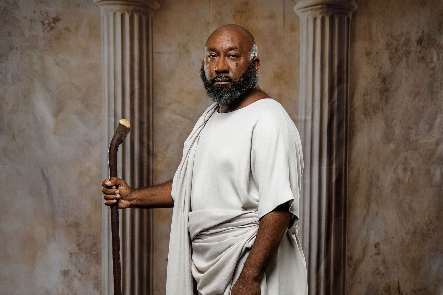 Black man in white ancient Greek toga holding wooden staff posing before marble columns in studio.