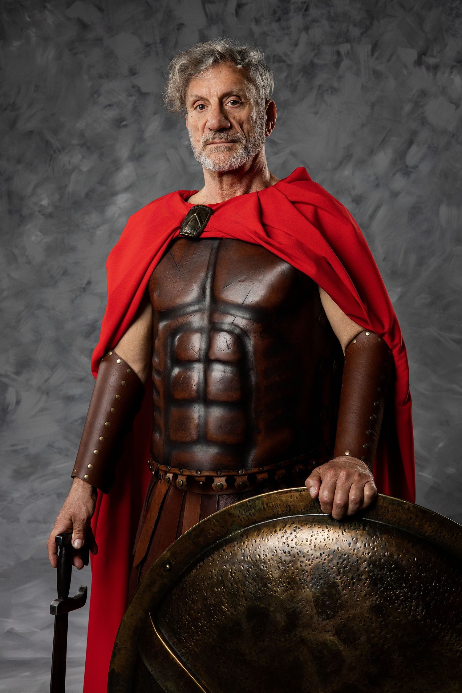Older man in Roman warrior costume with brown armor, red cape, holding sword and bronze shield against gray background.