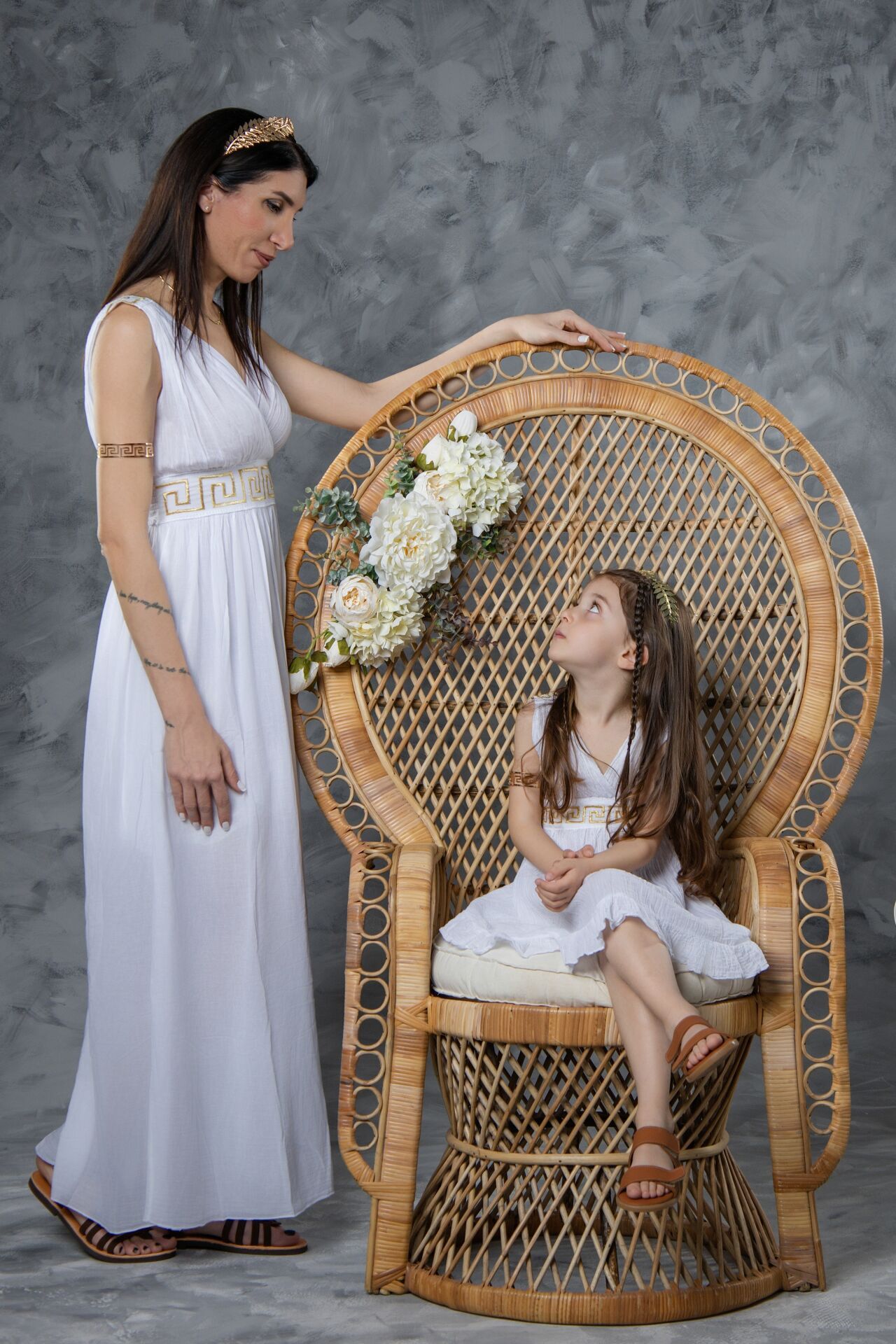 Woman in white Greek goddess gown stands beside girl in white dress seated in large wicker peacock chair with flowers.