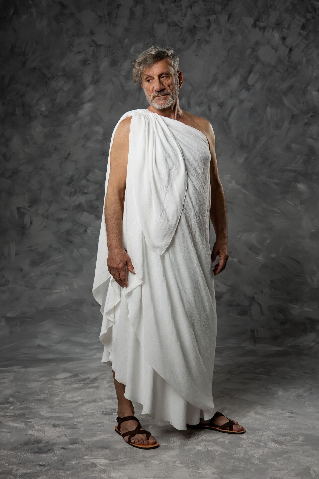 Older man in white Roman toga and sandals poses against gray textured studio background.
