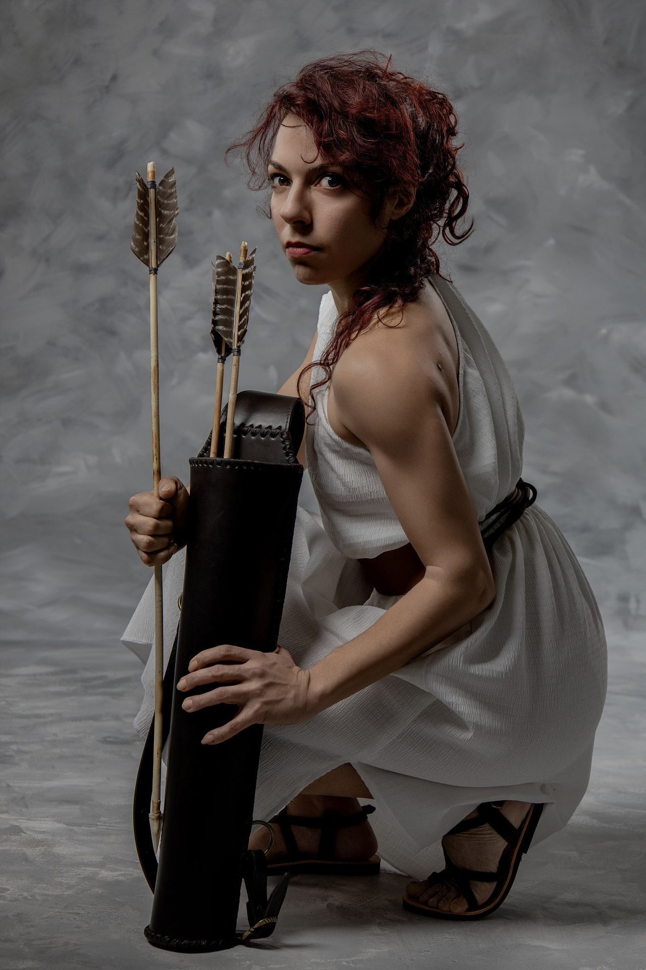A woman in a white Greek toga crouches holding arrows and a quiver, portraying the goddess Artemis.
