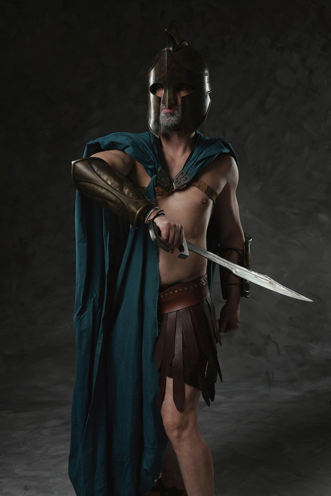 A figure in ancient warrior costume with leather armor and teal cape poses dramatically with a sword against dark background.