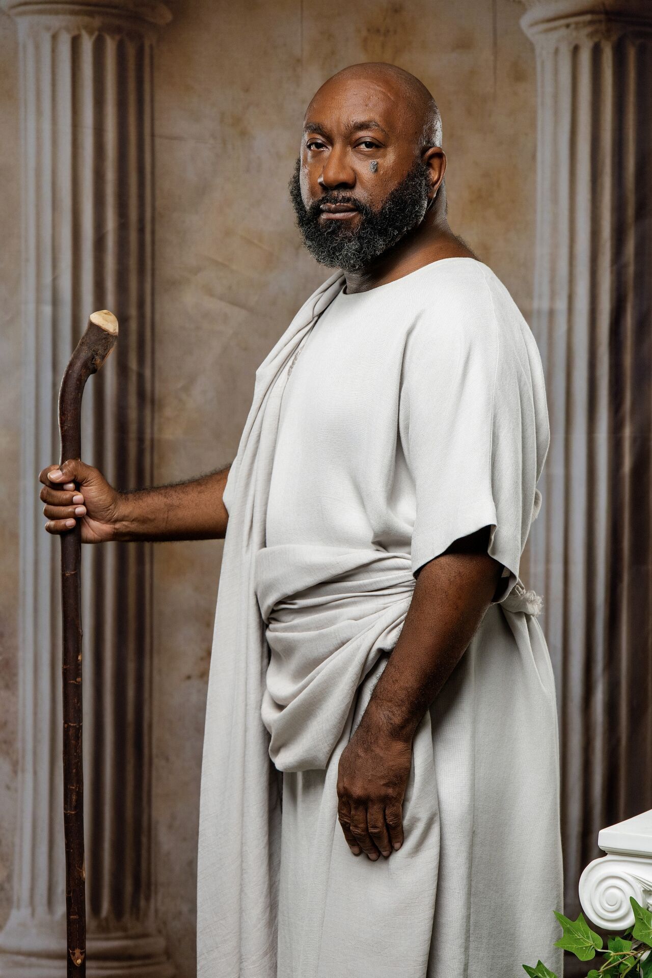 Elderly Black man in white Roman toga holding wooden staff, standing between stone columns in classical setting.