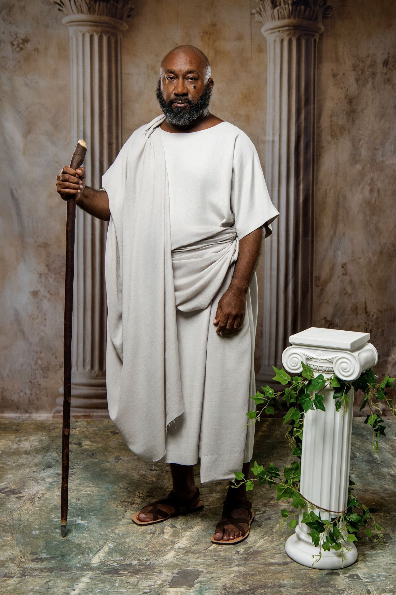 Man in ancient Roman toga holding a wooden staff, standing near a classical column with ivy, studio portrait.