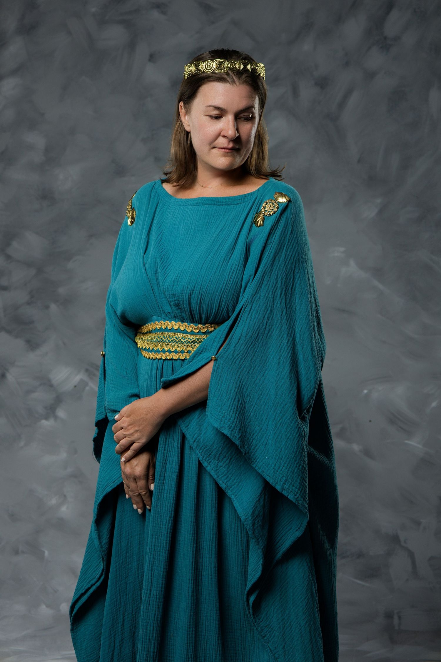 Woman in teal Greek goddess costume with gold crown and belt poses against gray background.