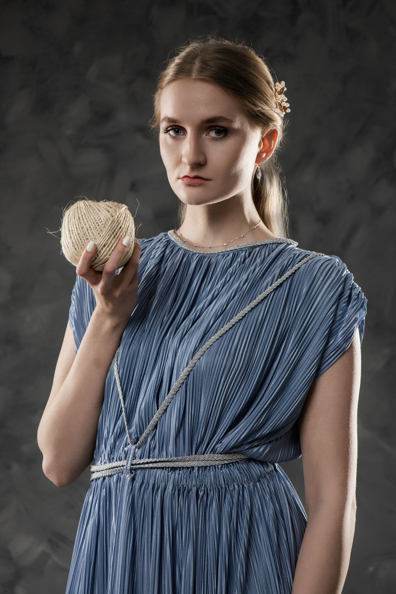 Young woman in blue pleated Greek-style dress holds twine ball, with rope belt and gold hair accessory, on gray background.