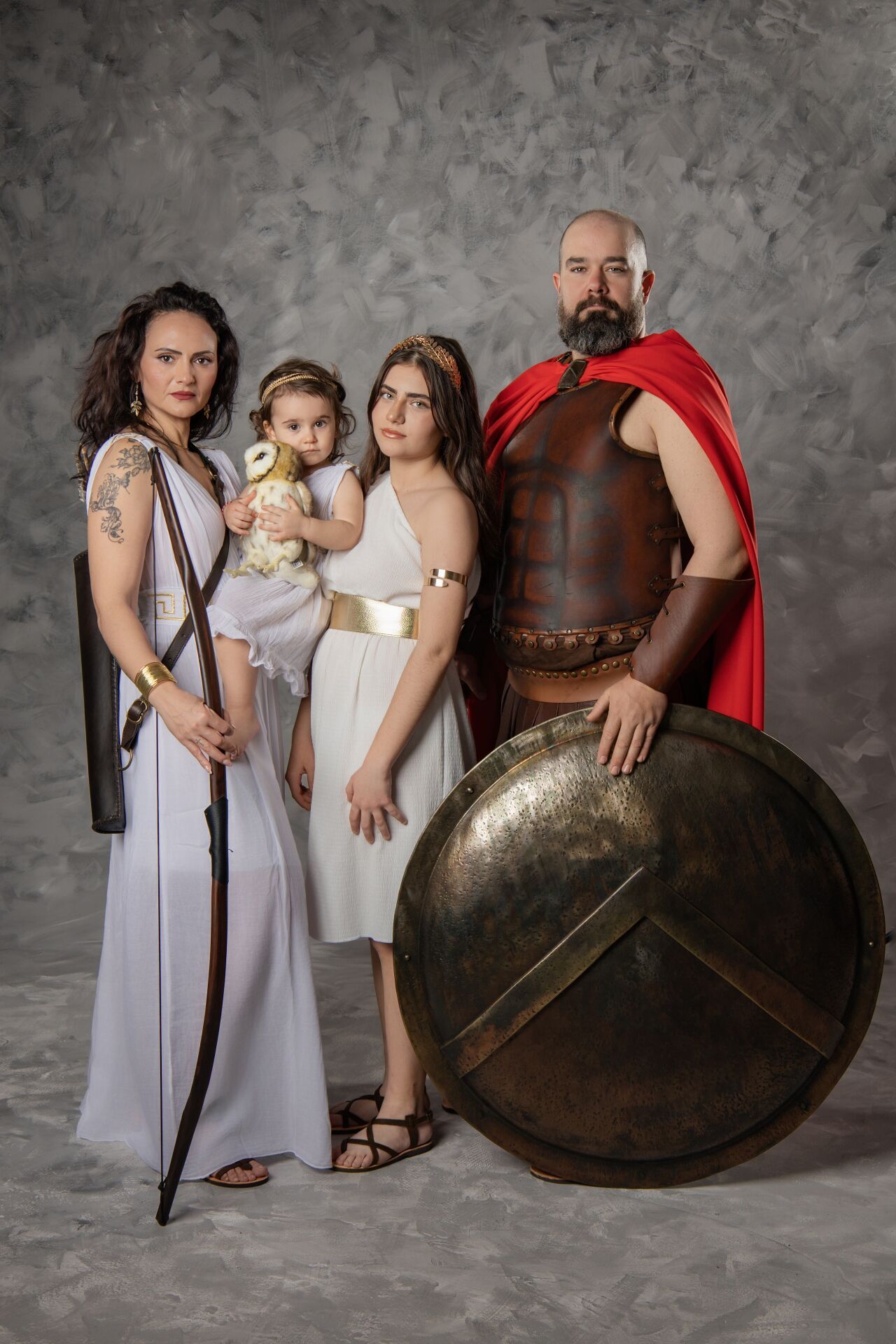 Family posing in ancient Greek and Spartan costumes, with man holding shield and red cape, women in white robes, and child.