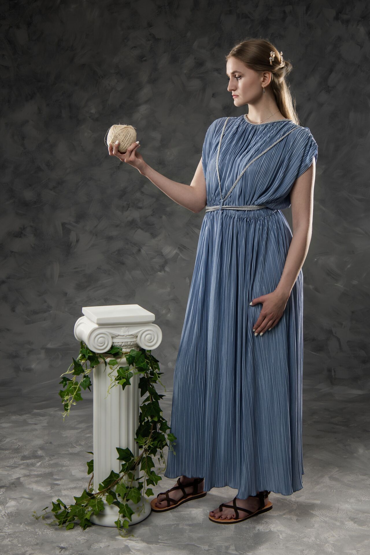Woman in blue Greek-style dress holding a stone sphere beside an ivy-draped white column on gray background.