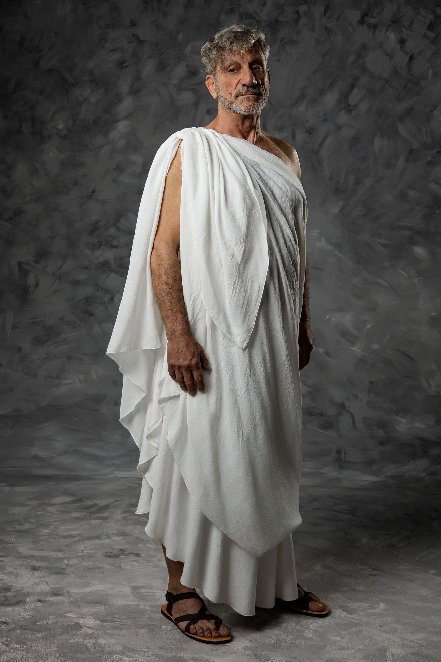 Older man in white Roman toga and sandals poses against gray backdrop.