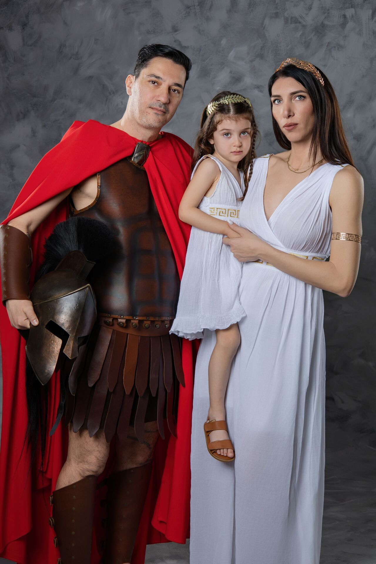 A family dressed in ancient Greek style costumes poses together against a gray backdrop for a themed portrait.