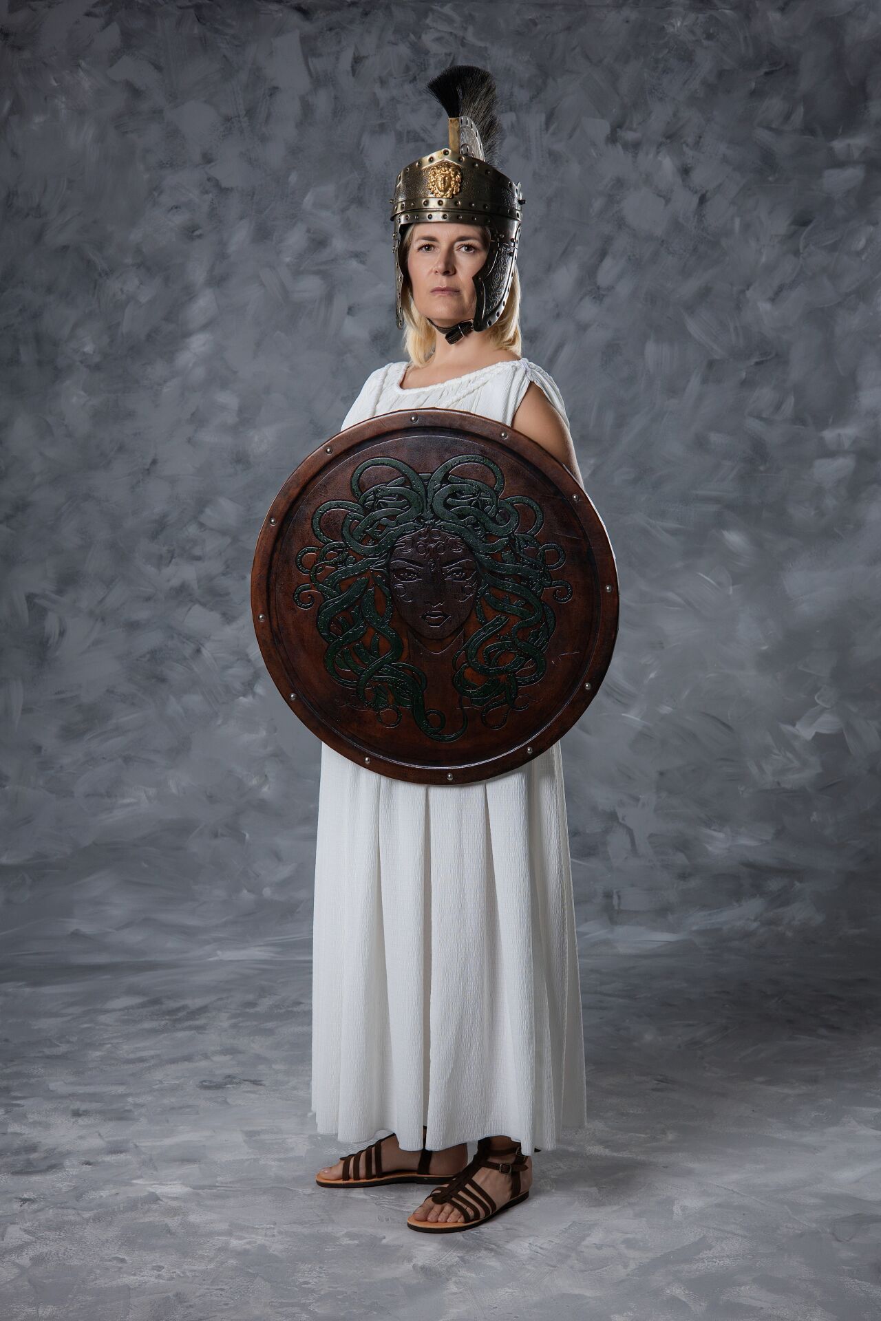 Woman dressed as Greek goddess Athena in white robe, golden helmet, and round shield with Medusa face on gray background.
