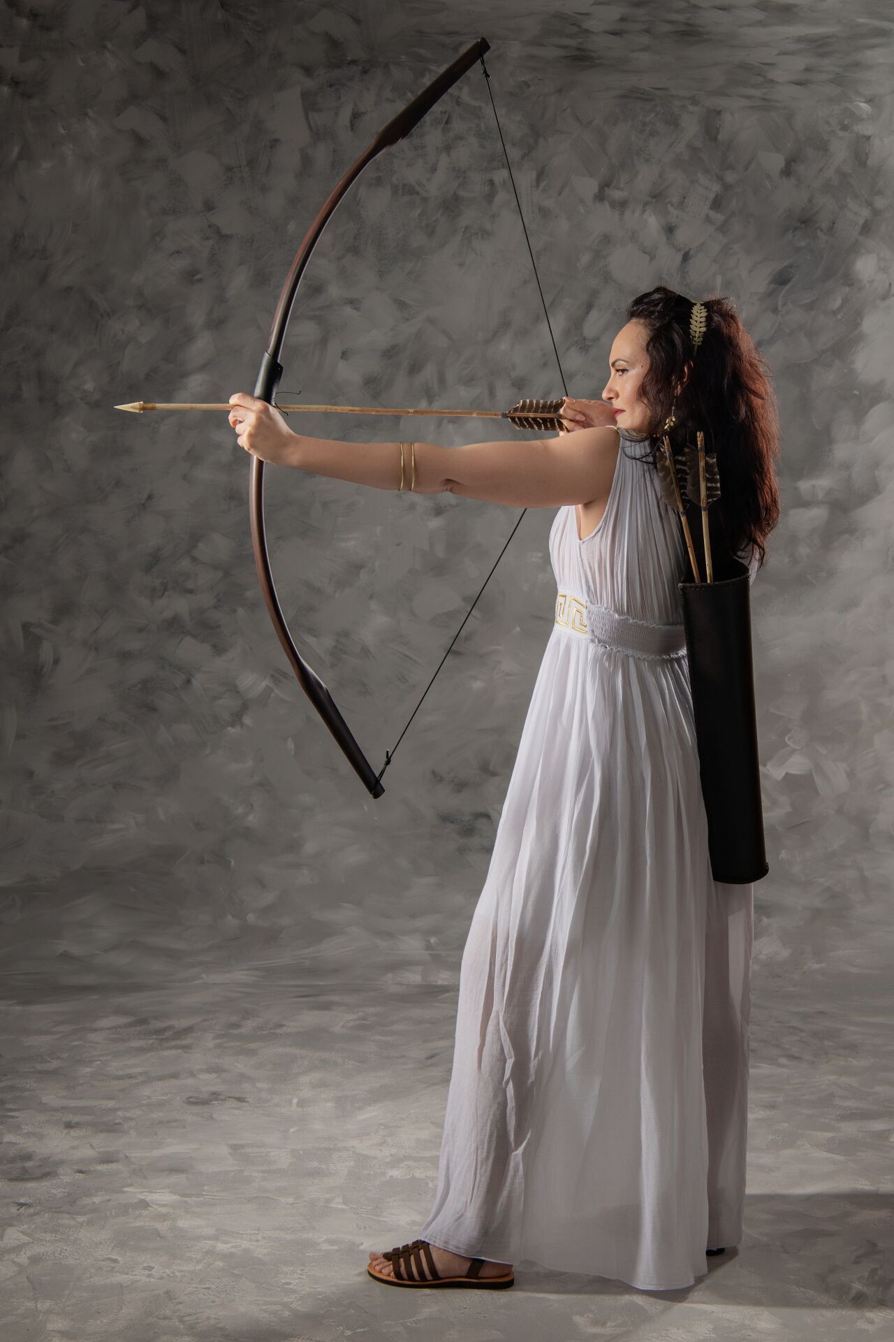 An archer in a flowing white dress draws a traditional wooden bow against a textured gray backdrop.