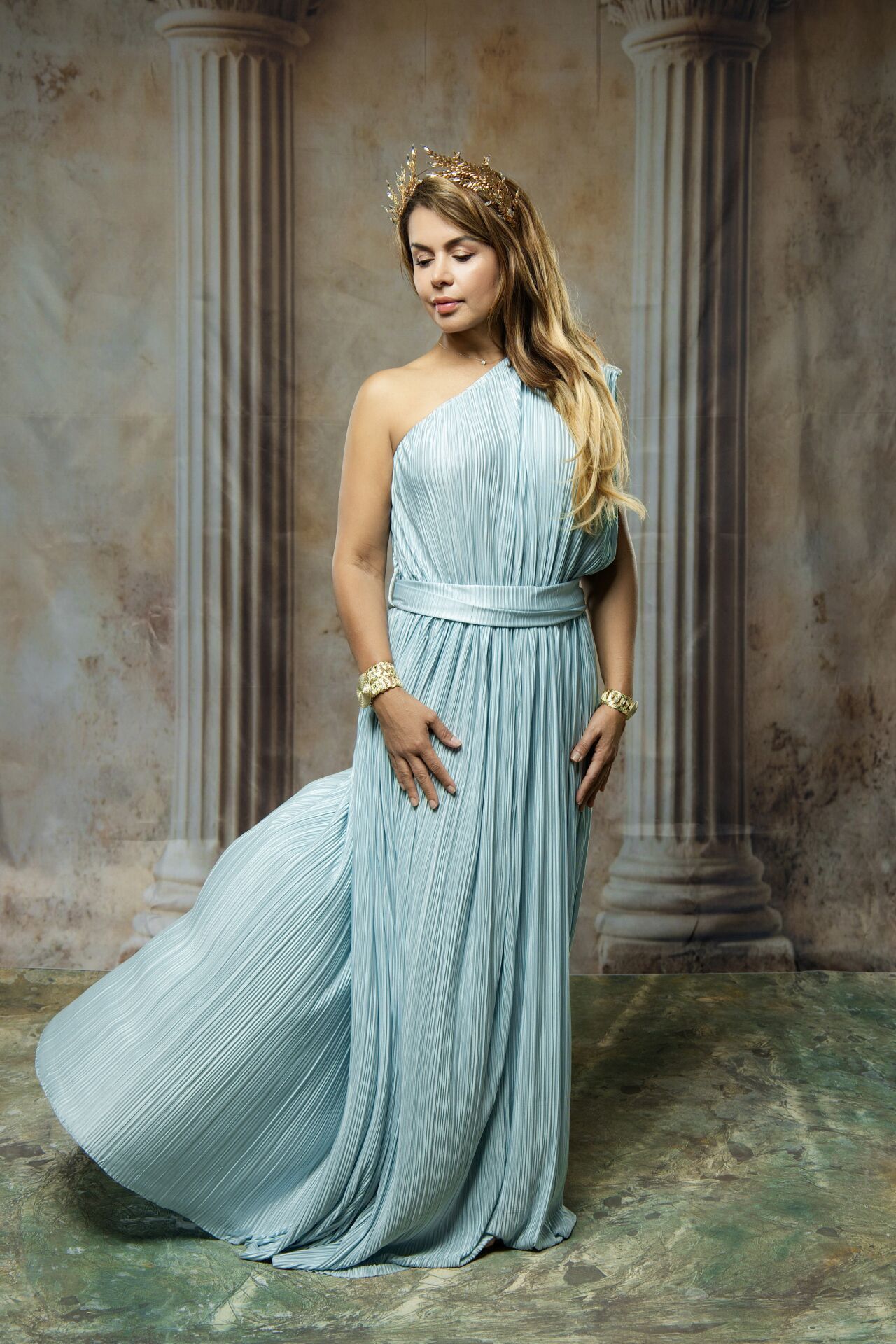 Woman in light blue pleated one-shoulder gown with gold crown and bracelet posing in elegant classical setting.