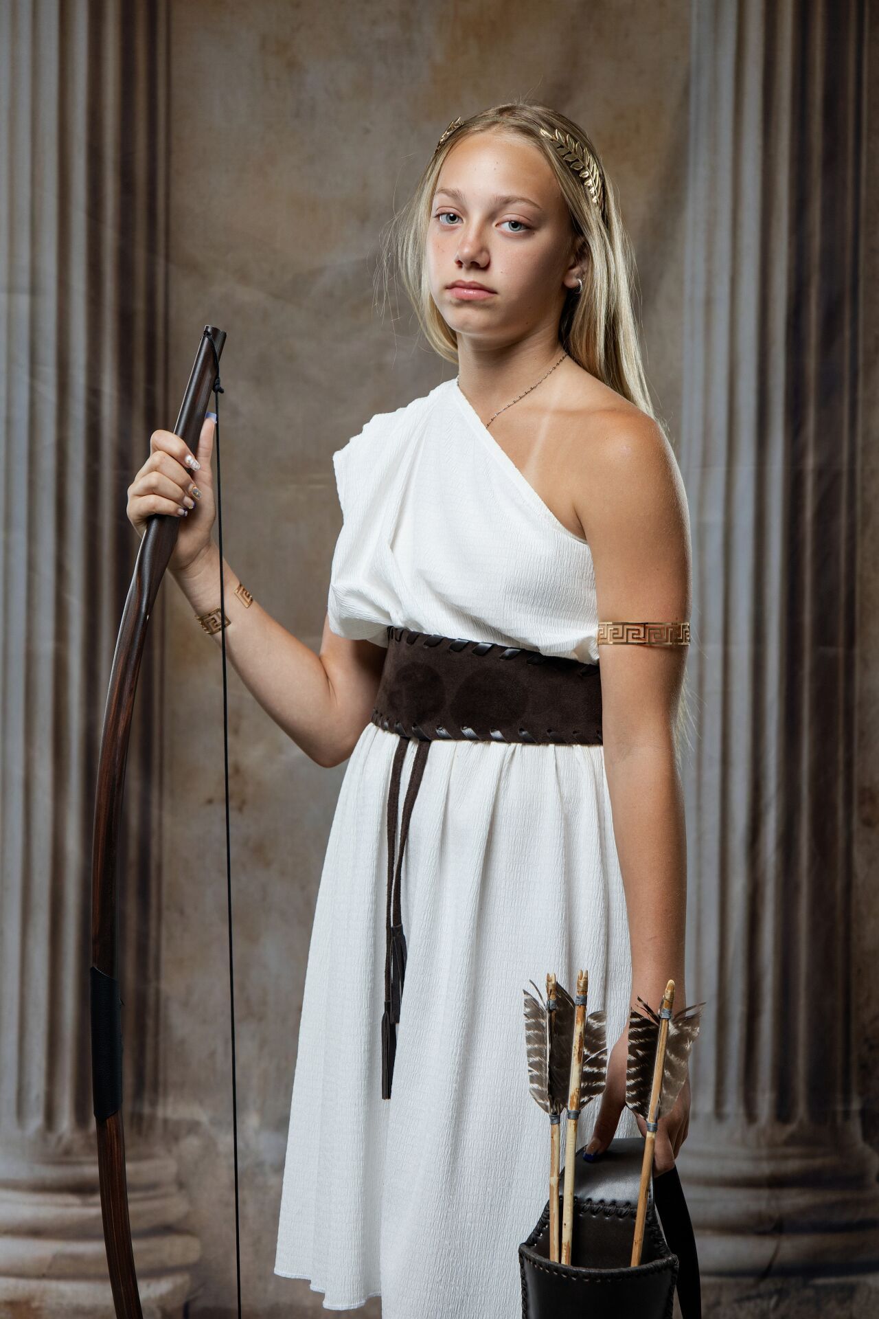 Blonde woman dressed as Greek goddess Artemis in white draped dress, holding bow with arrow quiver and gold accessories.