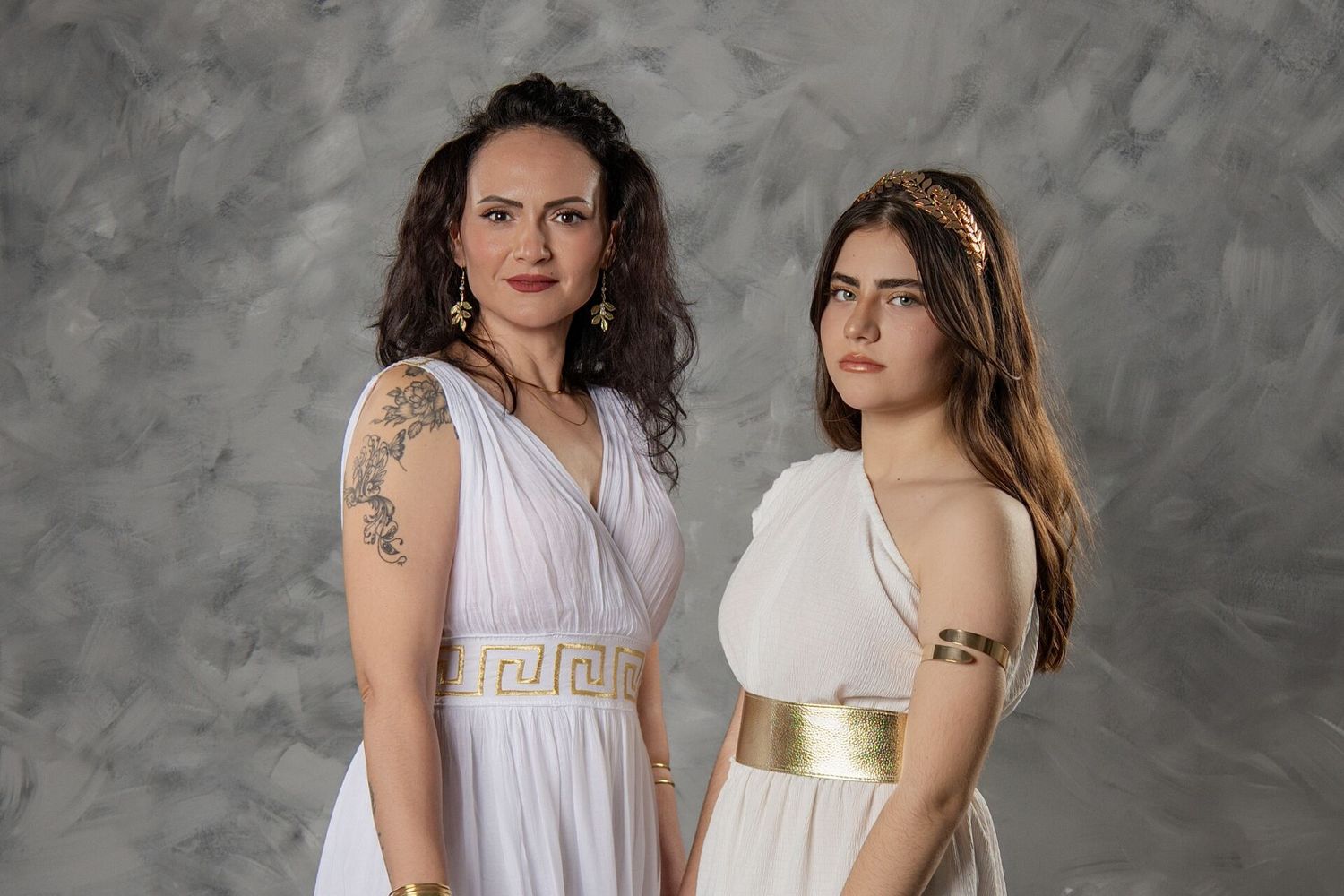 Two people in white outfits pose together against a textured gray background in a studio setting.