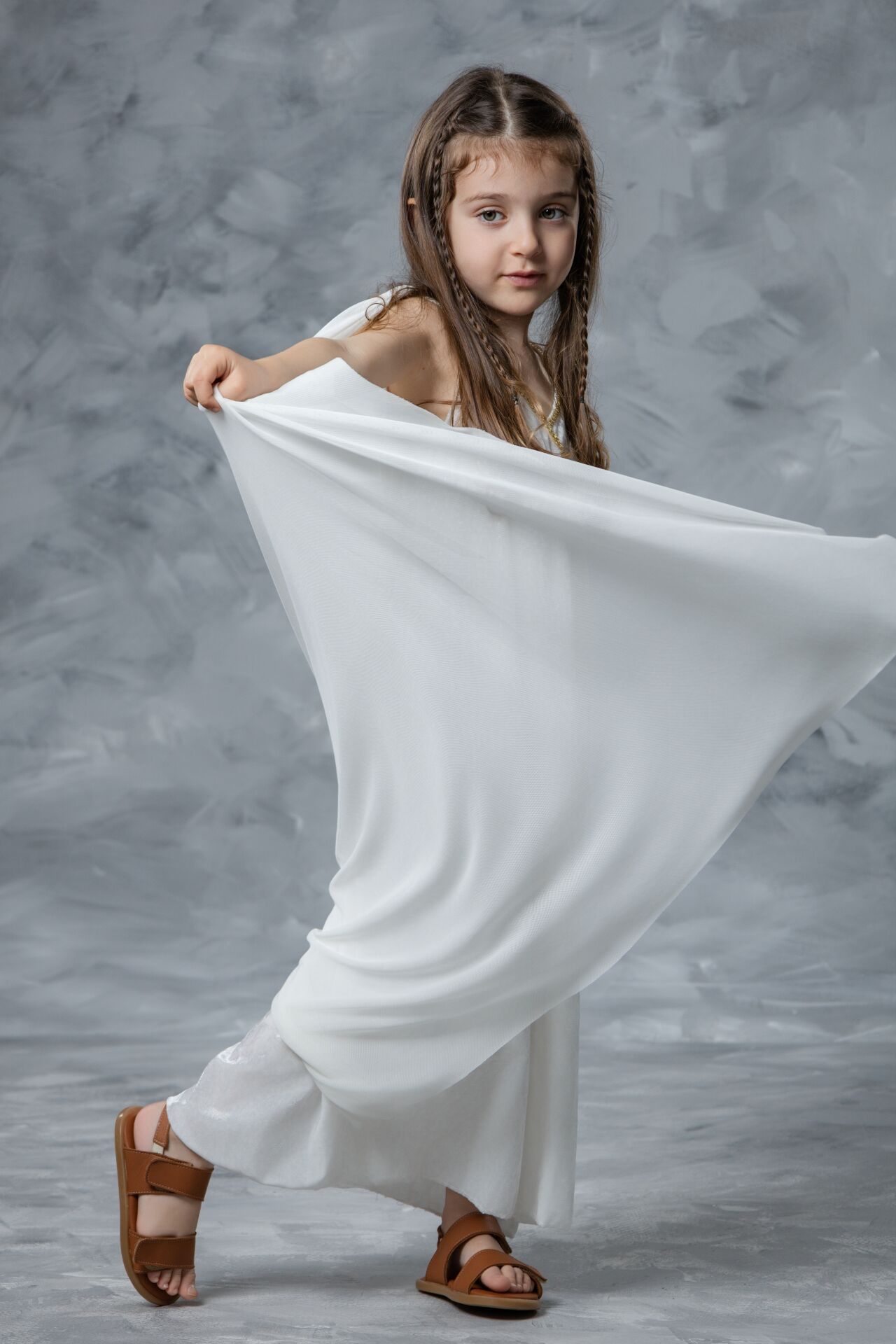 Young girl in flowing white toga poses gracefully with fabric against grey background.