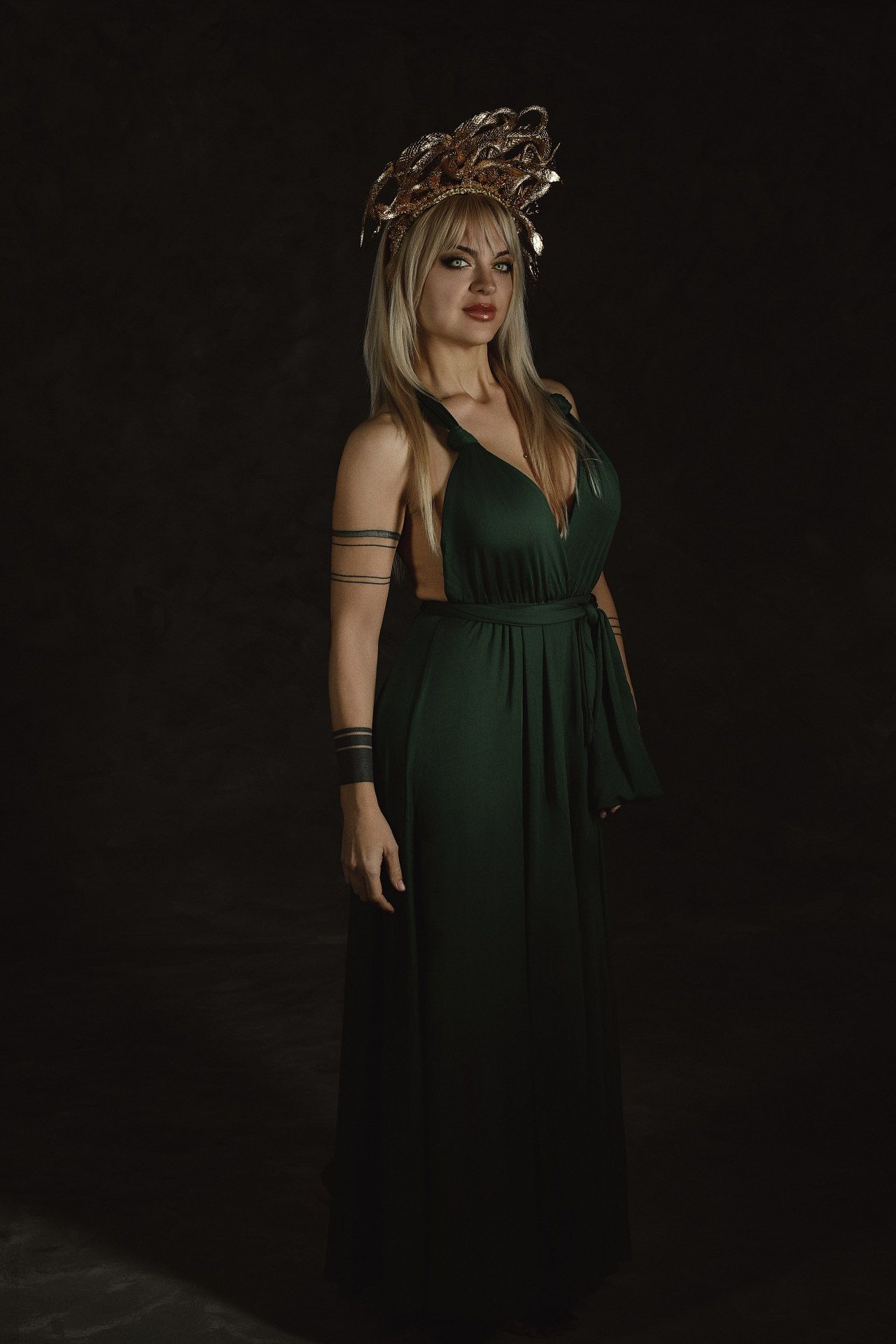 A figure in a dark green empire waist dress and floral crown poses against a black background in a moody portrait series.