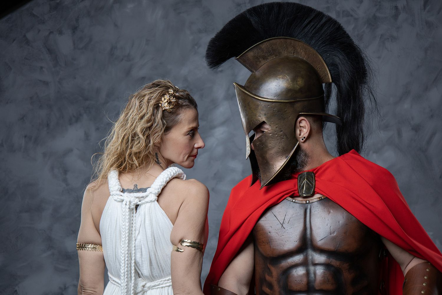 Woman in white Greek dress faces a muscular Spartan warrior in bronze helmet and red cape.