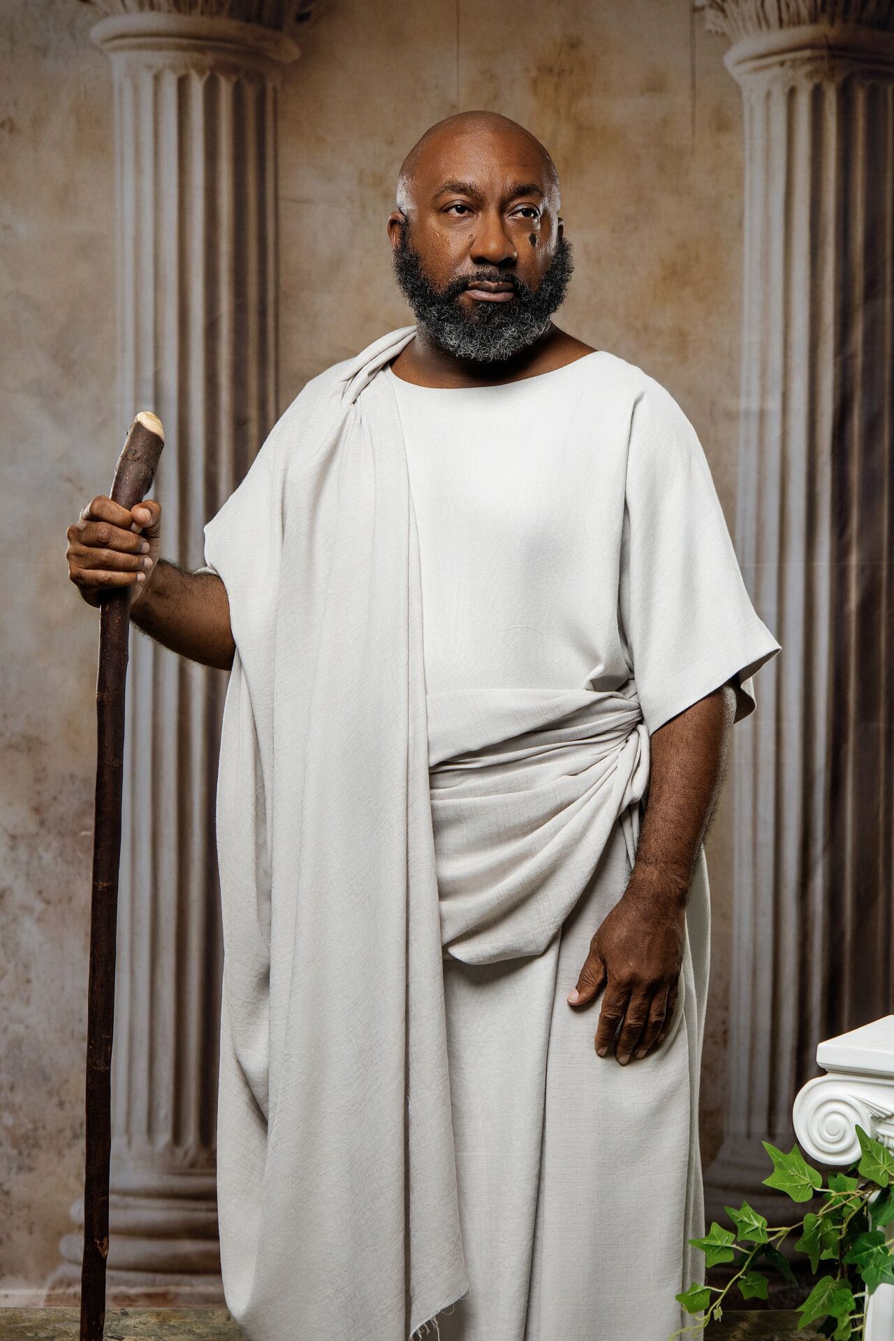 Bald Black man with gray beard in white Greek toga holding staff beside marble columns and ivy.