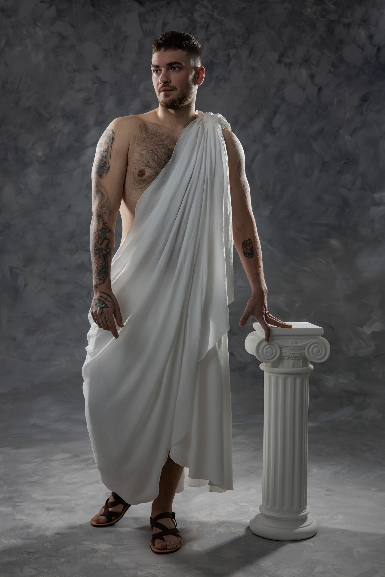 A person in a white toga and sandals poses dramatically next to a classical Roman column against a dark background.