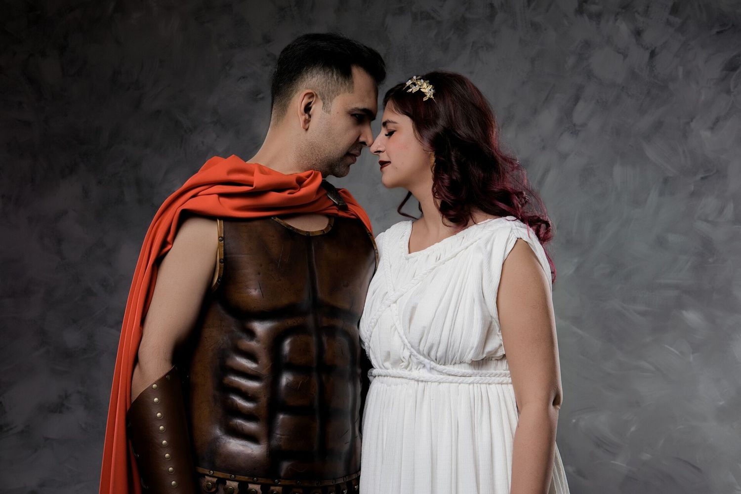 A man in Roman gladiator armor with red cape faces a woman in white Greek goddess dress and gold crown.