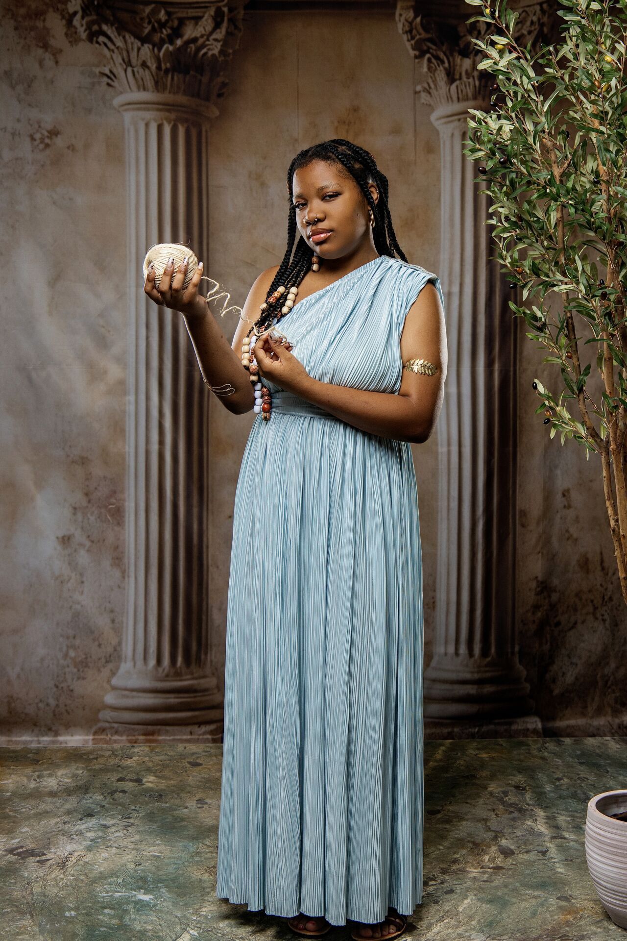 Woman in elegant blue pleated Grecian dress holds ornate sphere with beaded jewelry in studio.