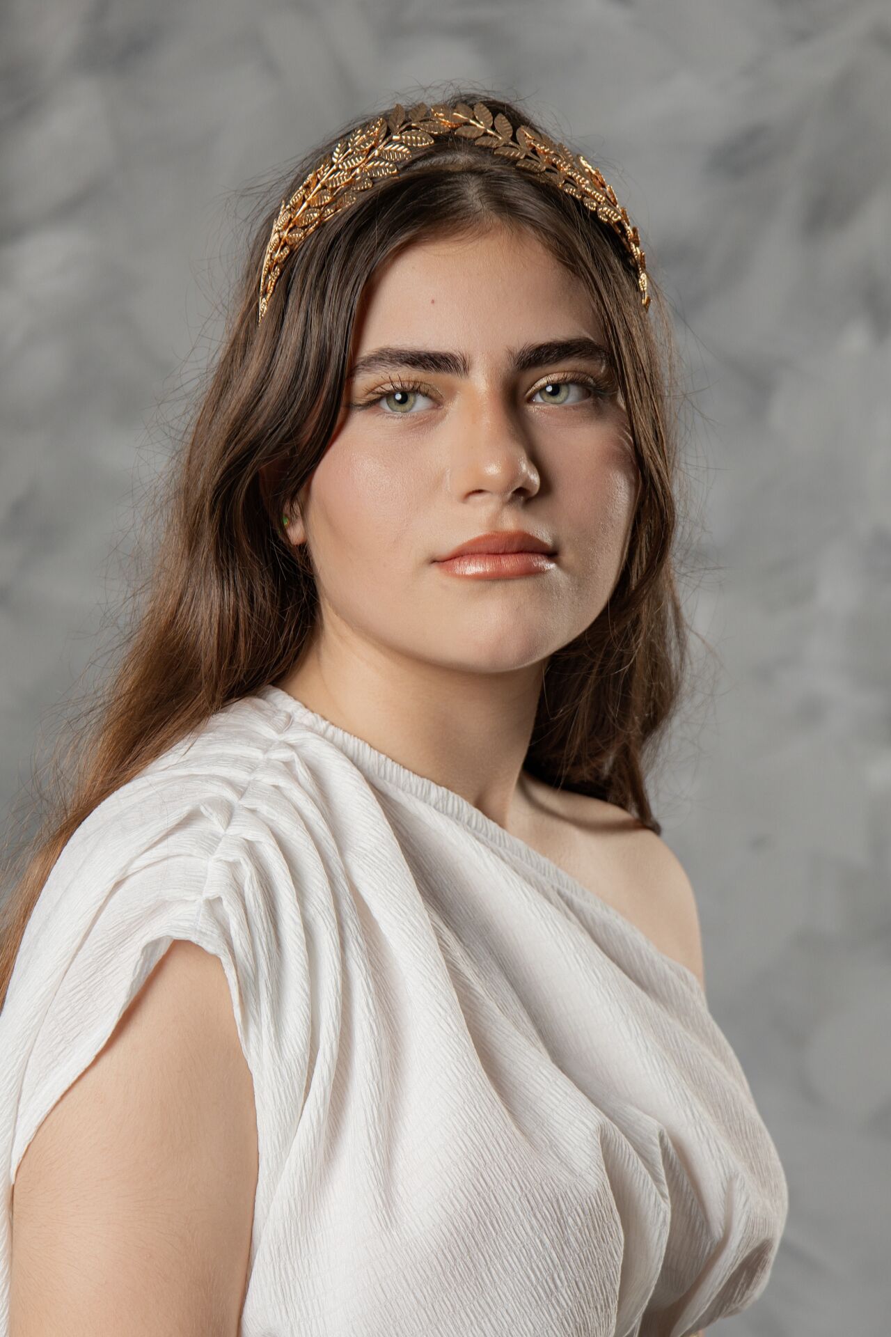 Portrait in a classical Greco-Roman style with white draped garment and gold leaf crown against a gray background.