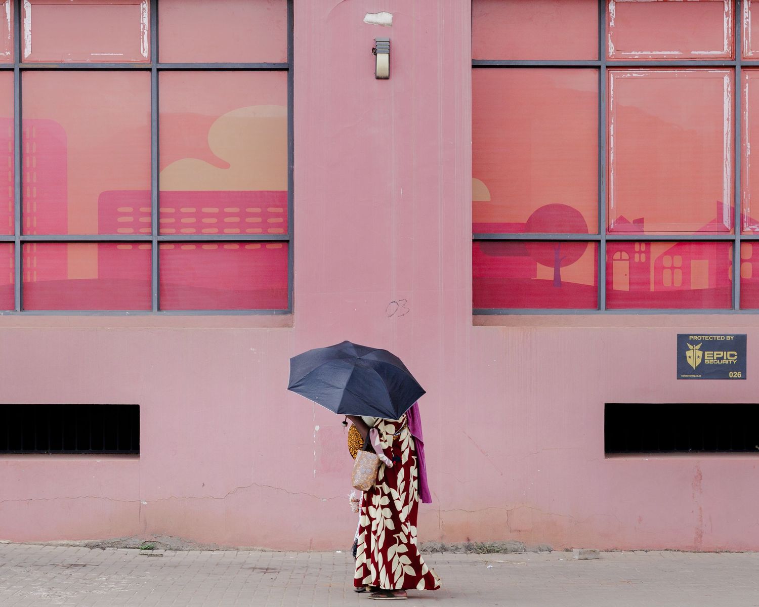 Minimalist street photography of a mother with an umbrella walking past a pink geometric building, exploring color and anonymity in Tanzania